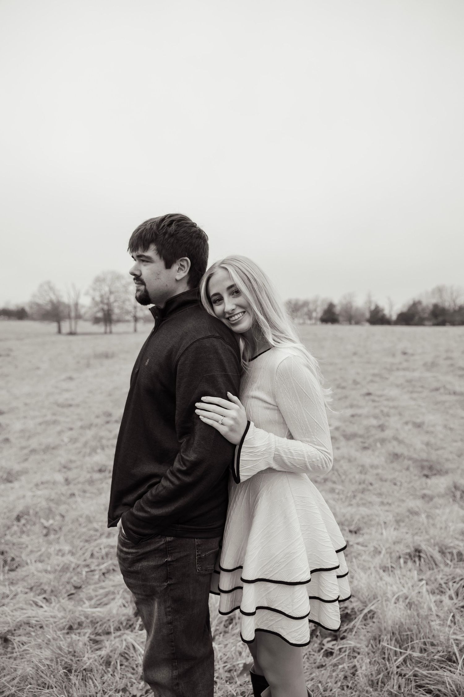 Black and white engagement photo of smiling blonde woman hugging man from behind in grassy field.
