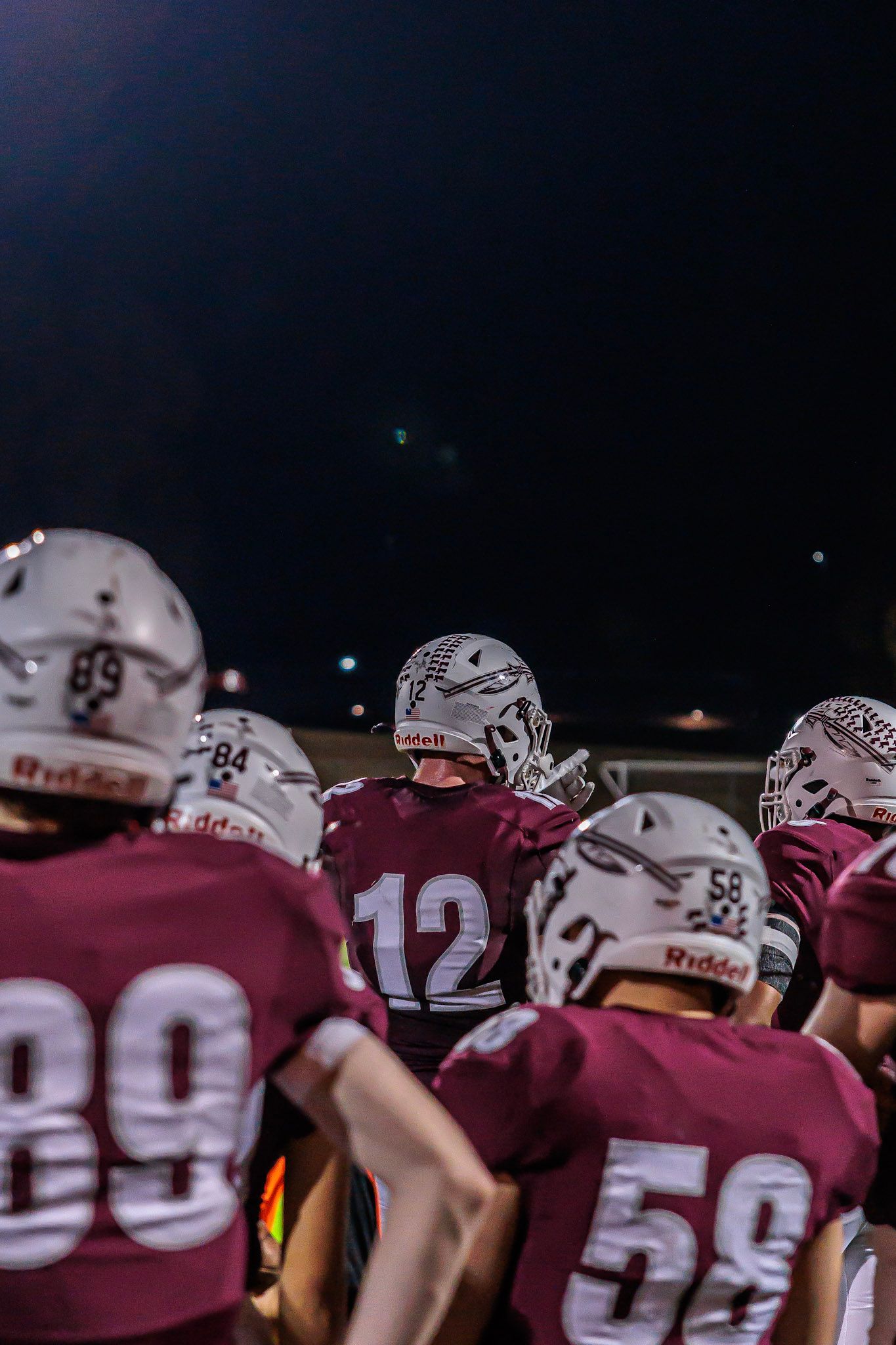 Football players in maroon uniforms and Riddell helmets huddling together under stadium lights at a night game.