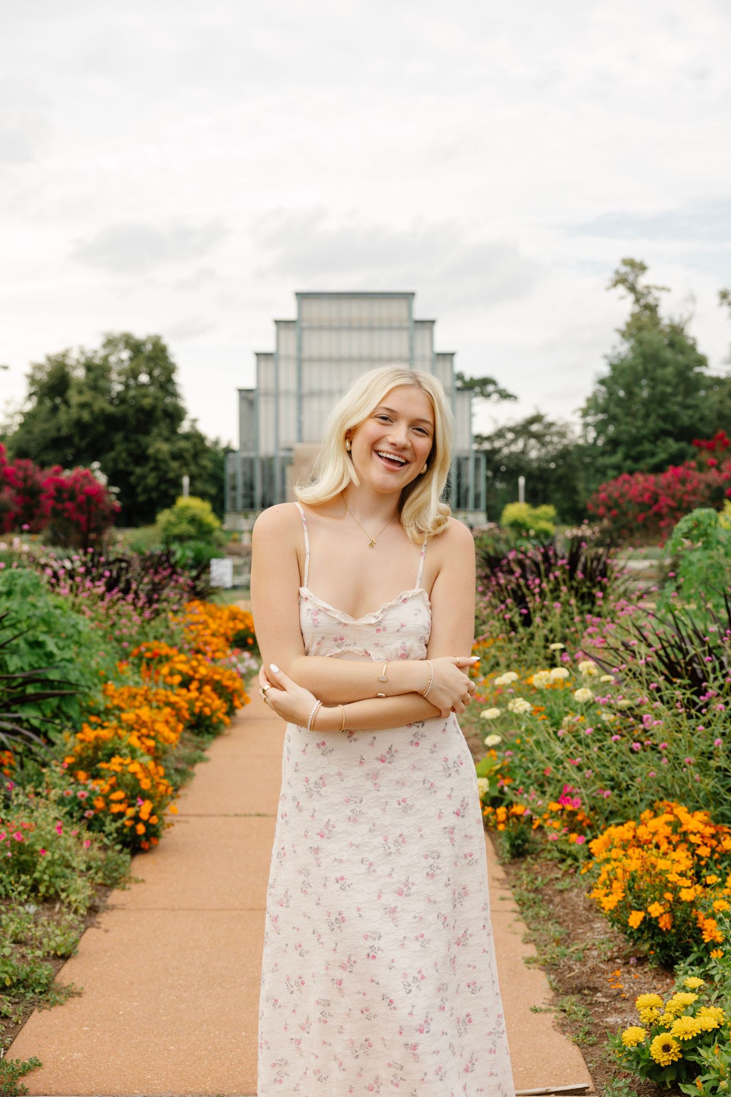 Smiling blonde woman in floral sundress standing in colorful garden path with greenhouse in background.