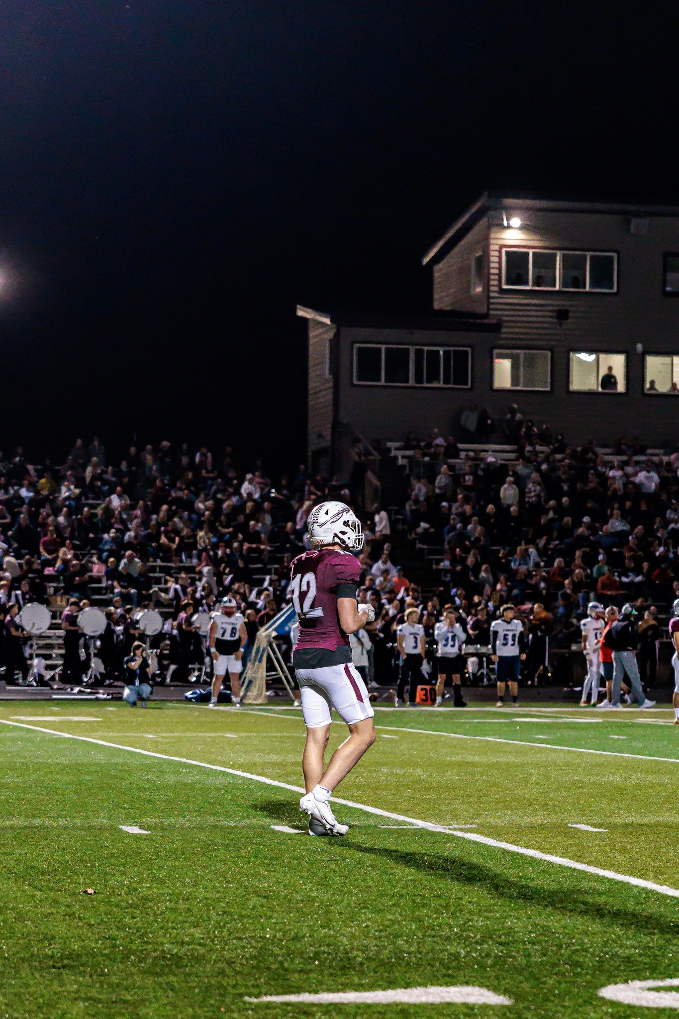High school football player #42 in maroon jersey stands on field during night game with crowd in stands.