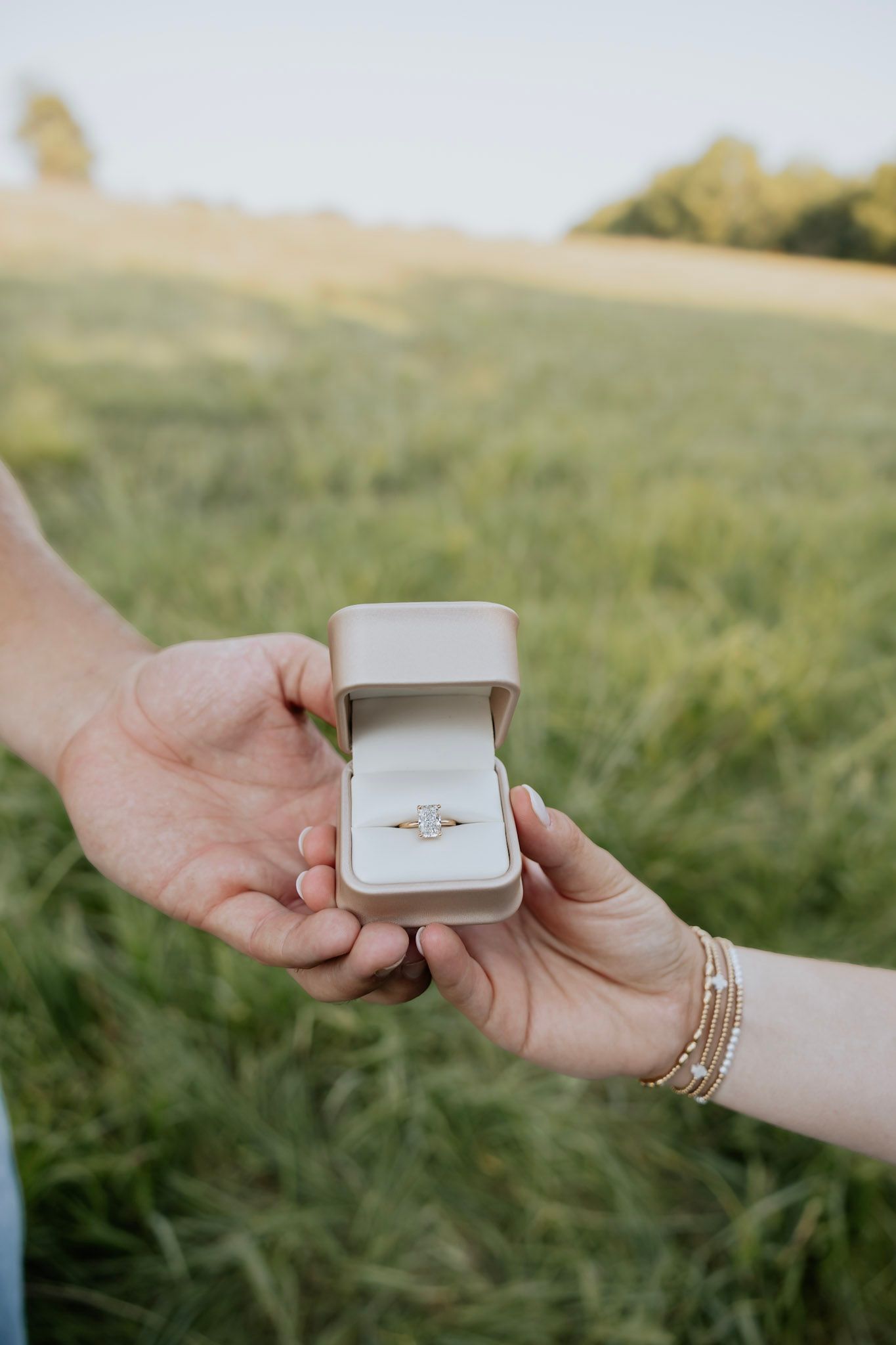 Close-up of couple holding open ring box with diamond engagement ring in grassy field.
