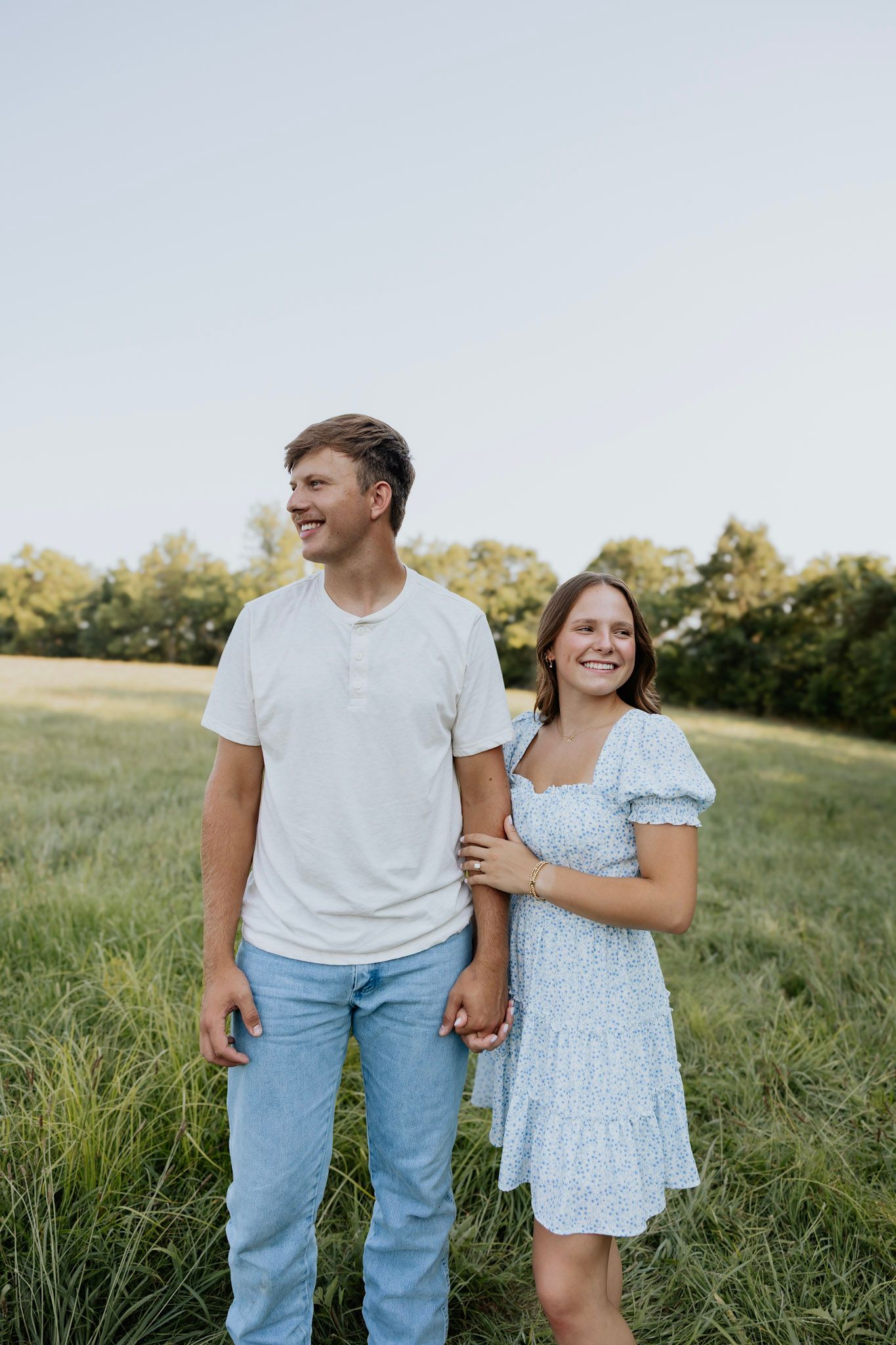Happy couple holding hands in a green field, woman in floral dress, man in white shirt and jeans.