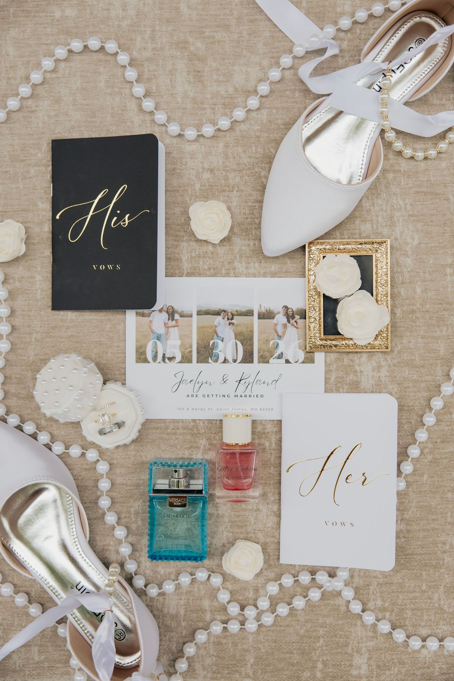 Flat lay of wedding details including vows books, invitation suite, pearl necklace, and white heels.