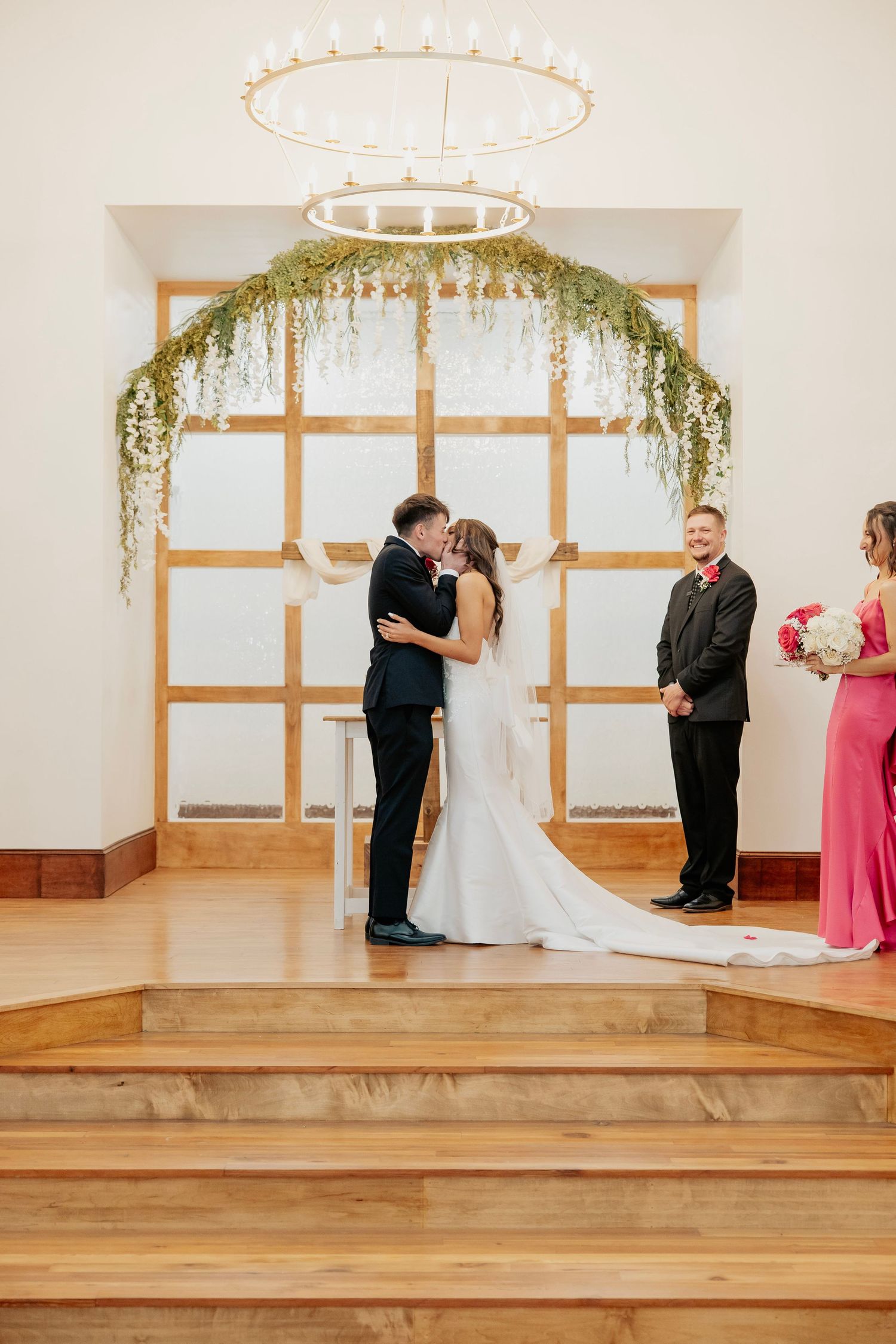 Bride and groom sharing first kiss at altar beneath floral arch in chapel.
