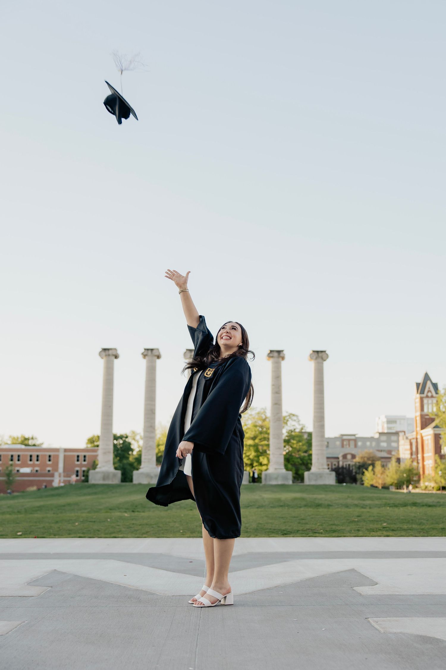 Female graduate in black gown tossing cap in the air near iconic university columns at golden hour.