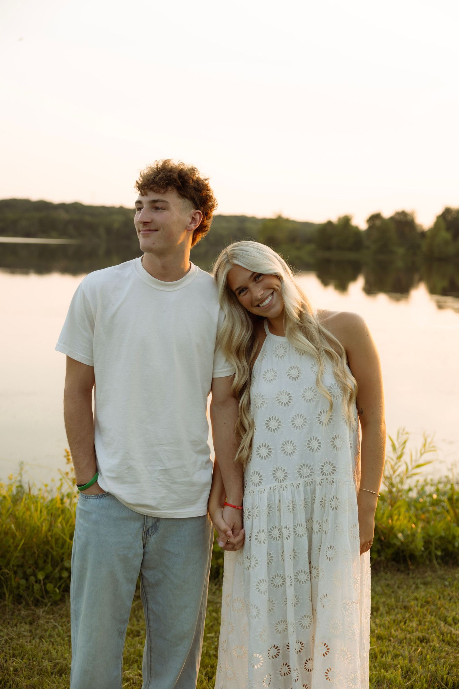 Young couple holding hands by a lake at sunset, woman in white dress smiling, man in white t-shirt and gray jeans.