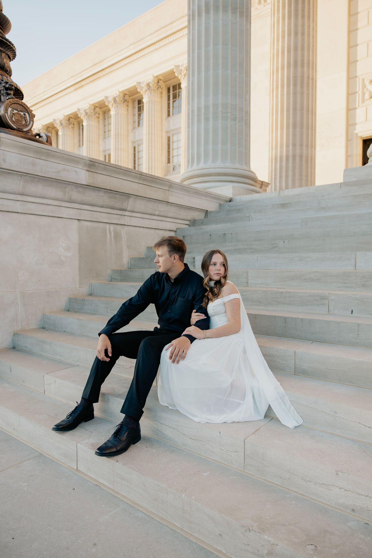 Bride in white dress and groom in black suit sitting on grand marble steps of a columned building.