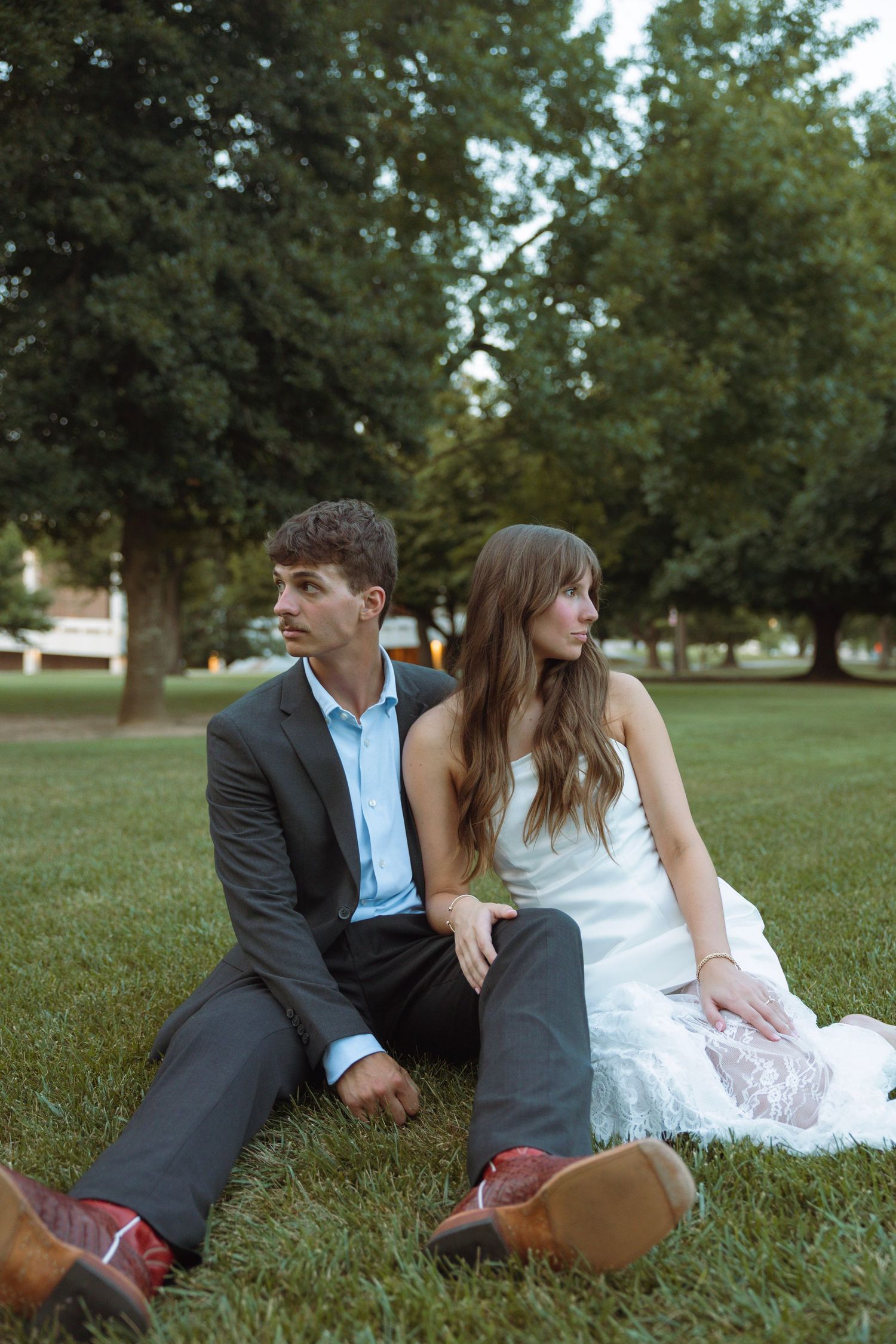 A young couple in wedding attire sits on green grass in a park, the groom in a suit and bride in a white lace dress.