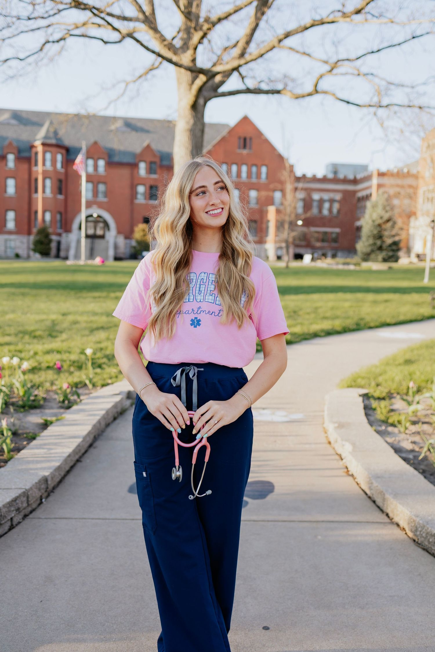 Blonde woman in pink nursing shirt and scrub pants standing on college campus pathway.