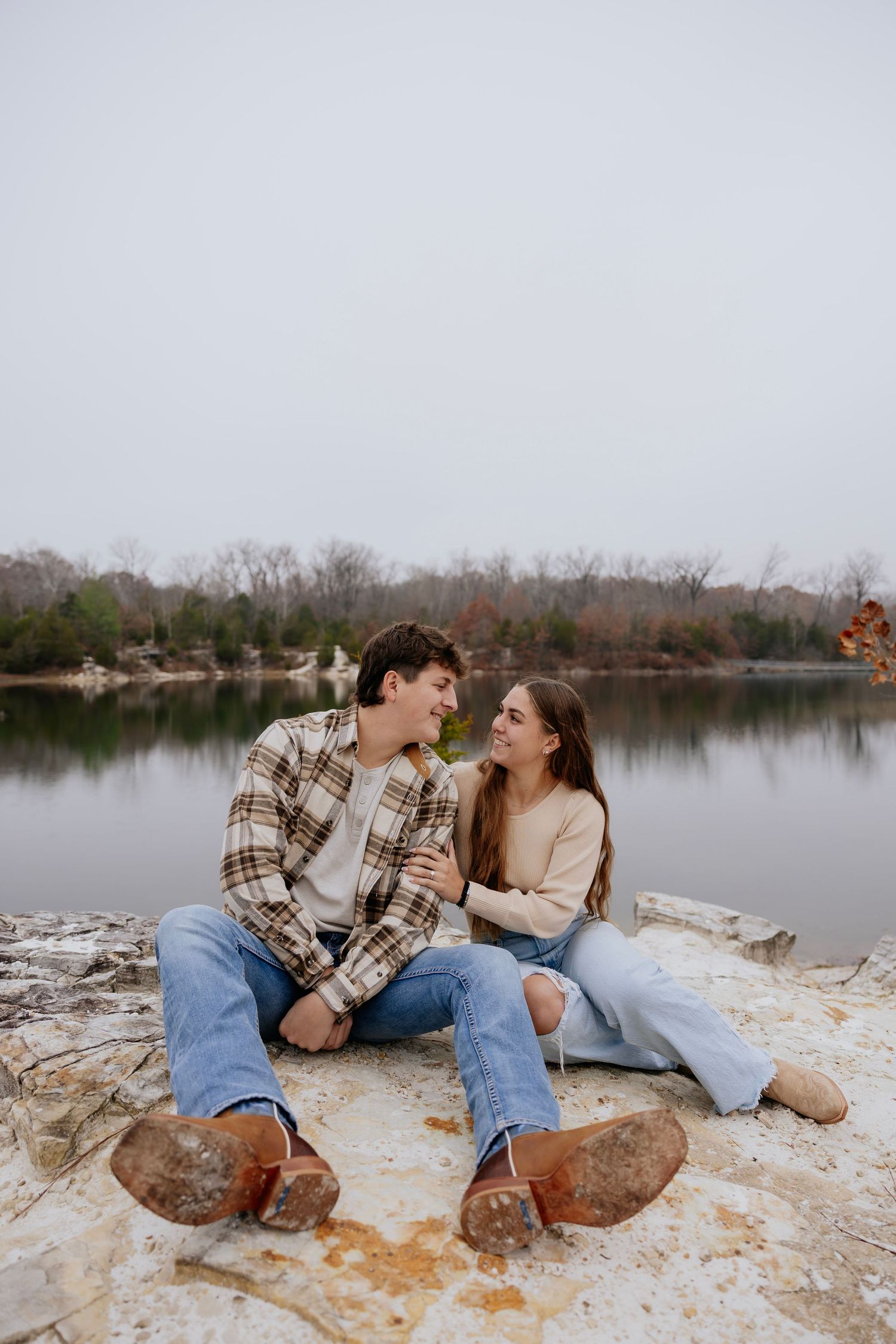 Happy couple sitting on rocks by a lake, smiling at each other on overcast day.