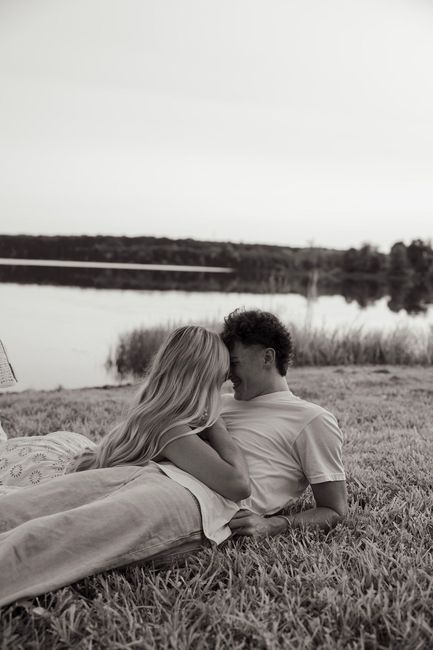 Black and white photo of a couple lying on grass by a lake, embracing romantically at dusk.