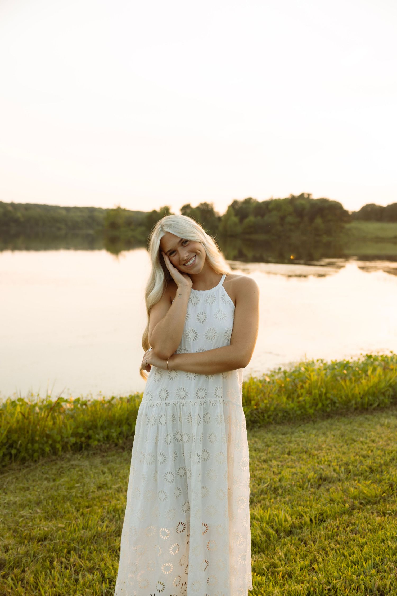 Smiling blonde woman in white eyelet maxi dress standing by a serene lake at golden hour sunset.