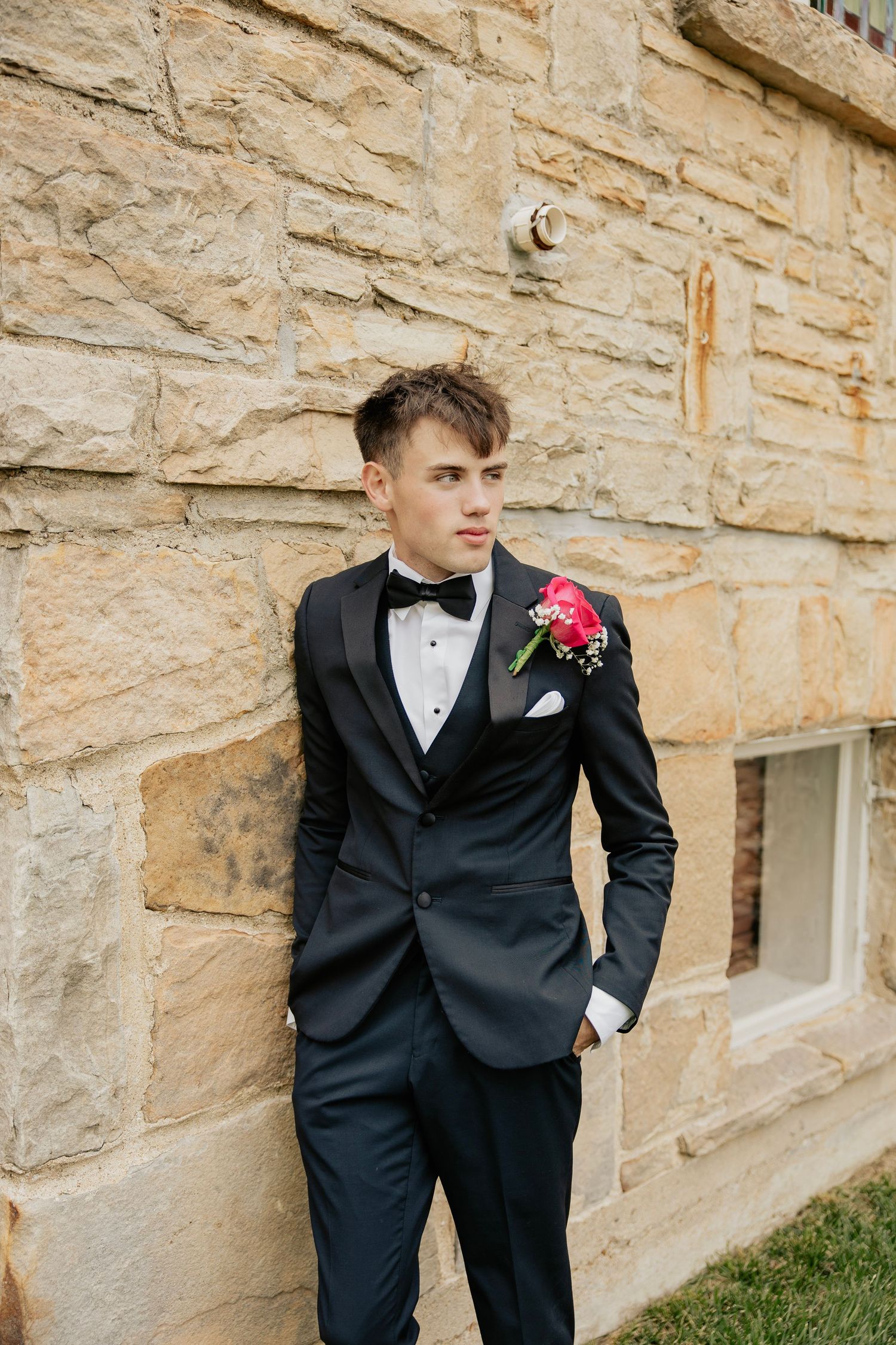 Handsome groom in black tuxedo with pink boutonniere leaning against rustic stone church wall.