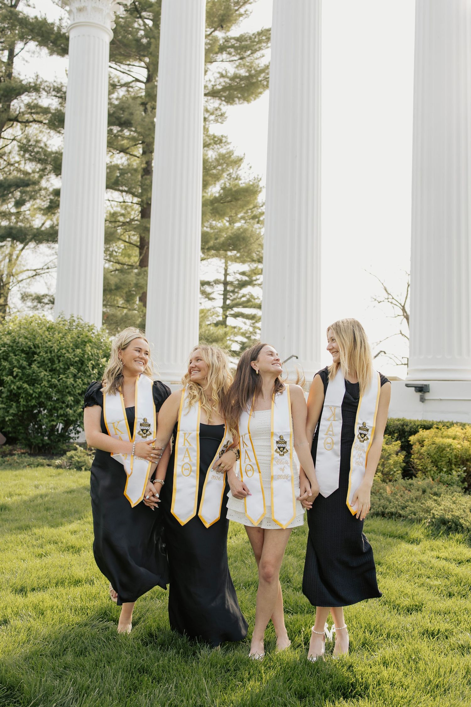 Four female graduates in black gowns with gold stoles laughing near white columns outdoors.