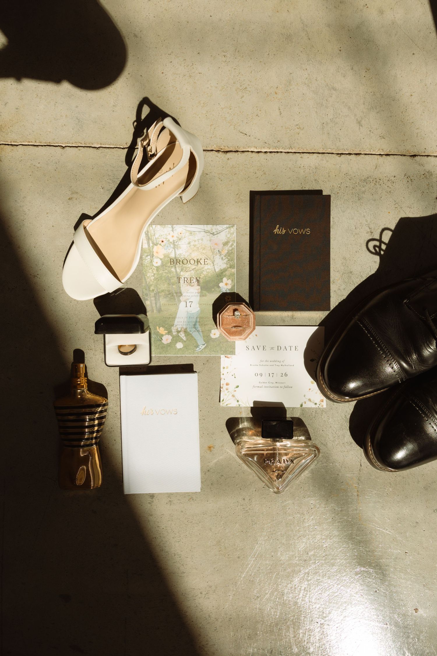 Wedding flat lay with bridal heels, groom dress shoes, save the date, vow books, rings, and perfume on concrete floor.