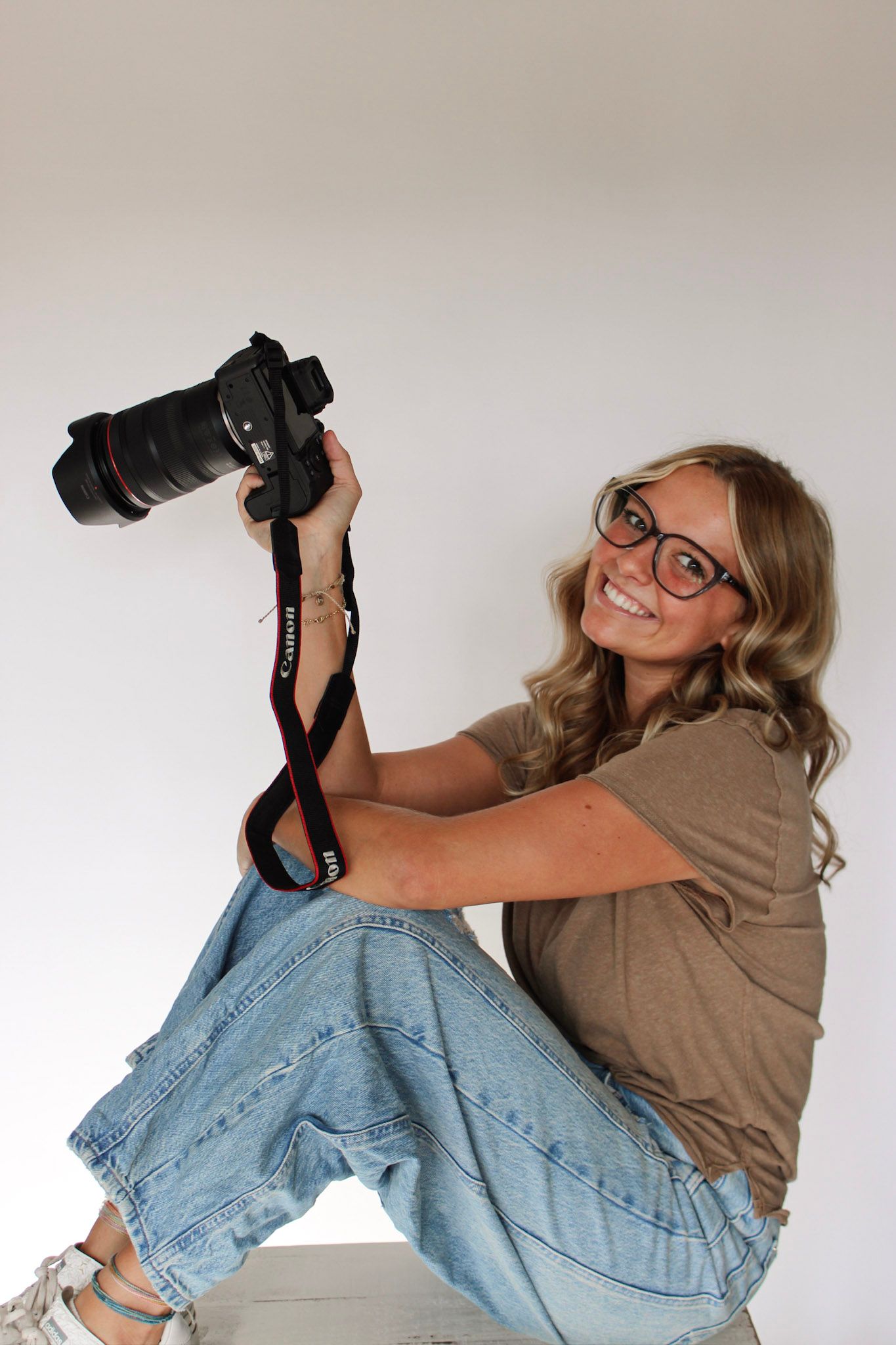 Cheerful blonde woman with glasses posing with a Canon DSLR camera on white background.