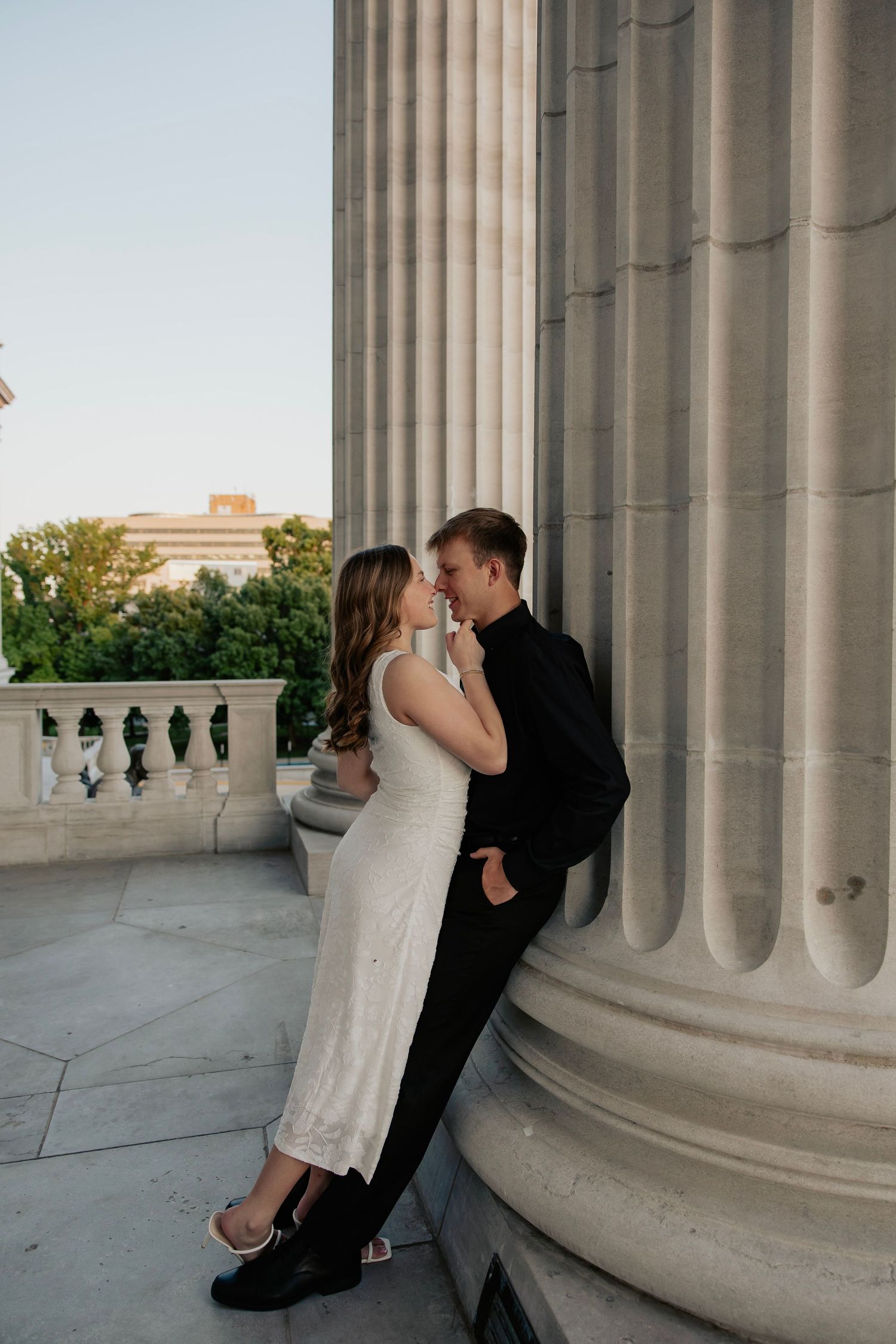 Bride and groom sharing an intimate moment leaning against a large stone column of a grand building.