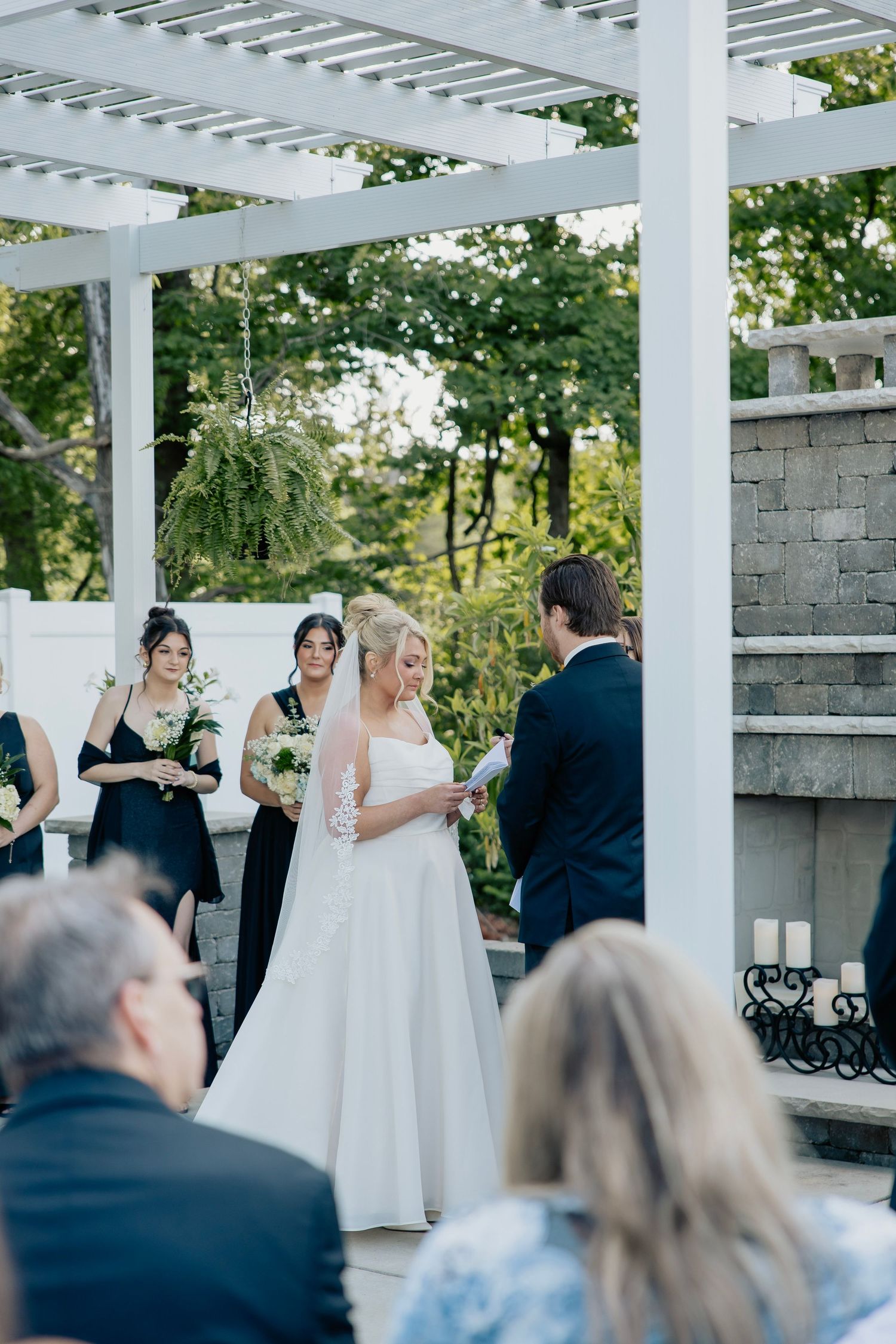Bride reads vows to groom under white pergola with bridesmaids in black dresses during outdoor wedding ceremony.