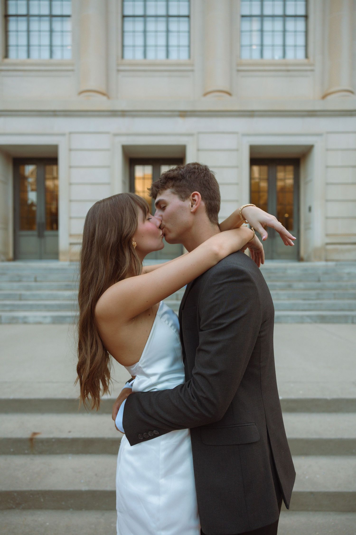 A couple shares a romantic kiss in front of a classical building, with the woman in a white dress and man in a dark suit.