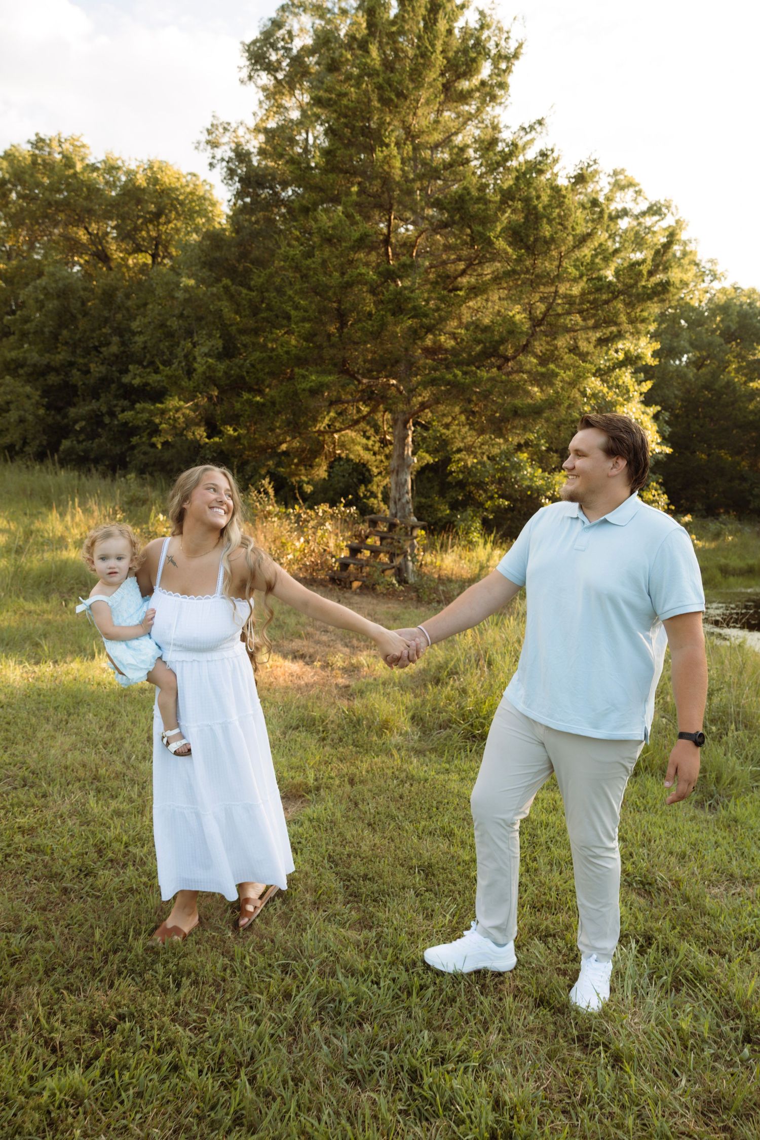 Happy young family holding hands in a sunny green field, mother in white dress carrying toddler.