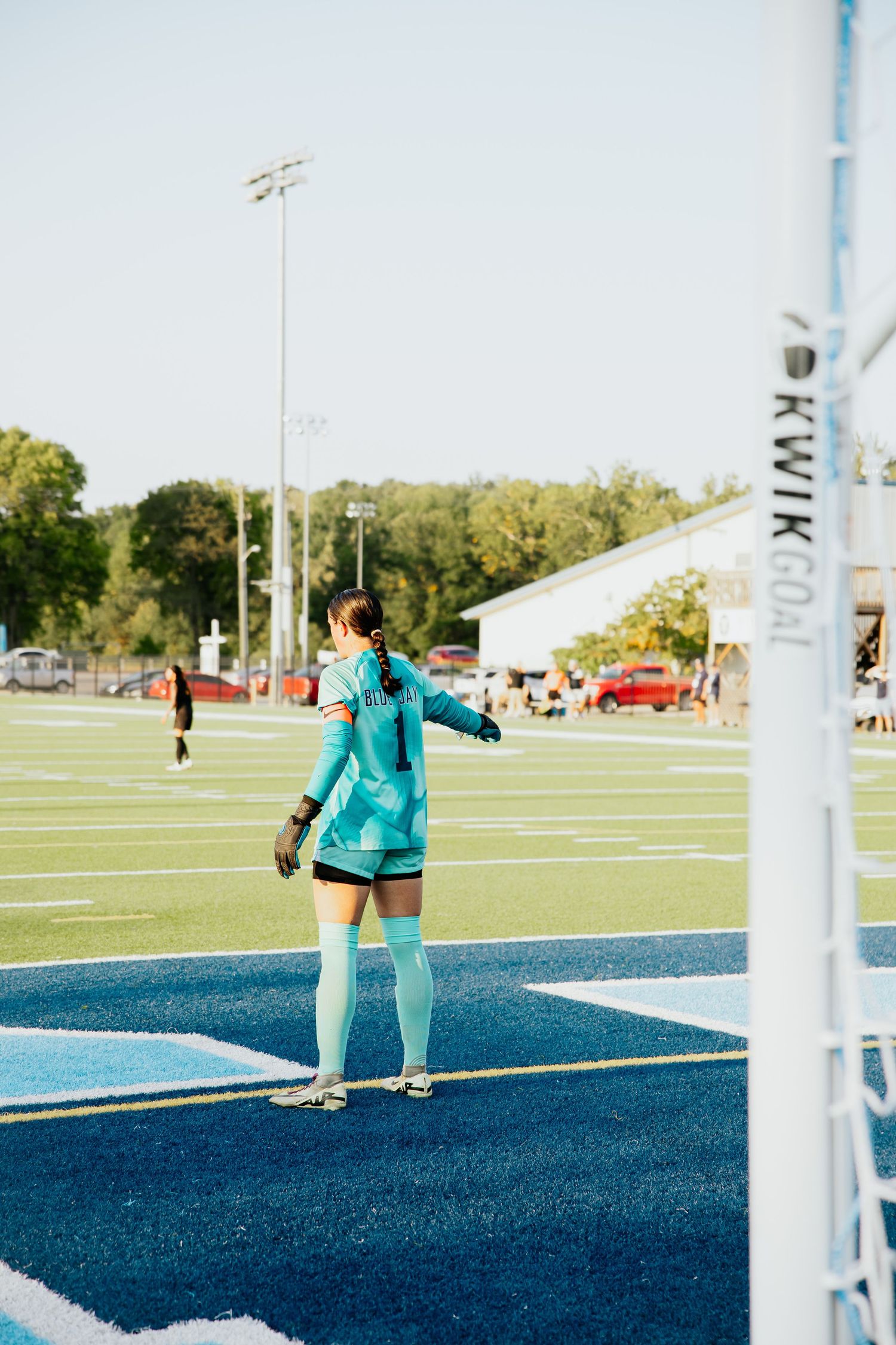 Female soccer goalkeeper in teal uniform stands near goal post on blue turf field during daytime game.