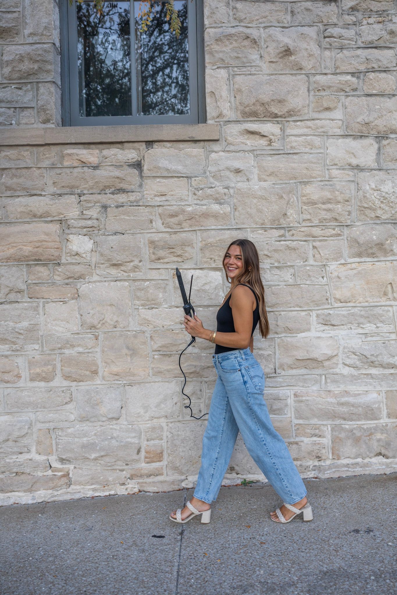 Cheerful young woman holding flat iron hair straightener, wearing black bodysuit and jeans, stone wall background.