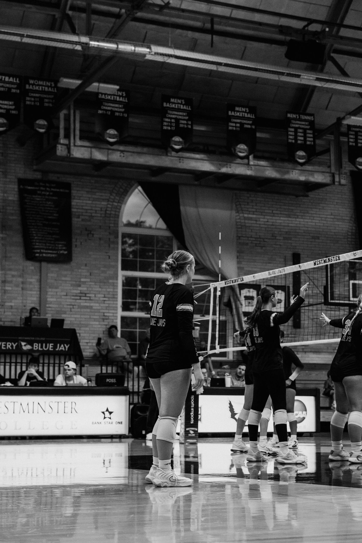 Black and white photo of female volleyball player #12 serving during indoor gym match at Westminster.