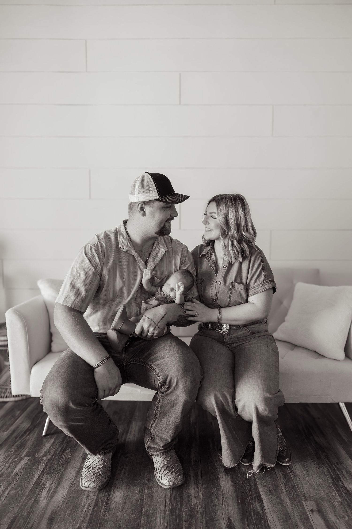 Black and white photo of a couple sitting on a couch, smiling at each other while holding a small dog.