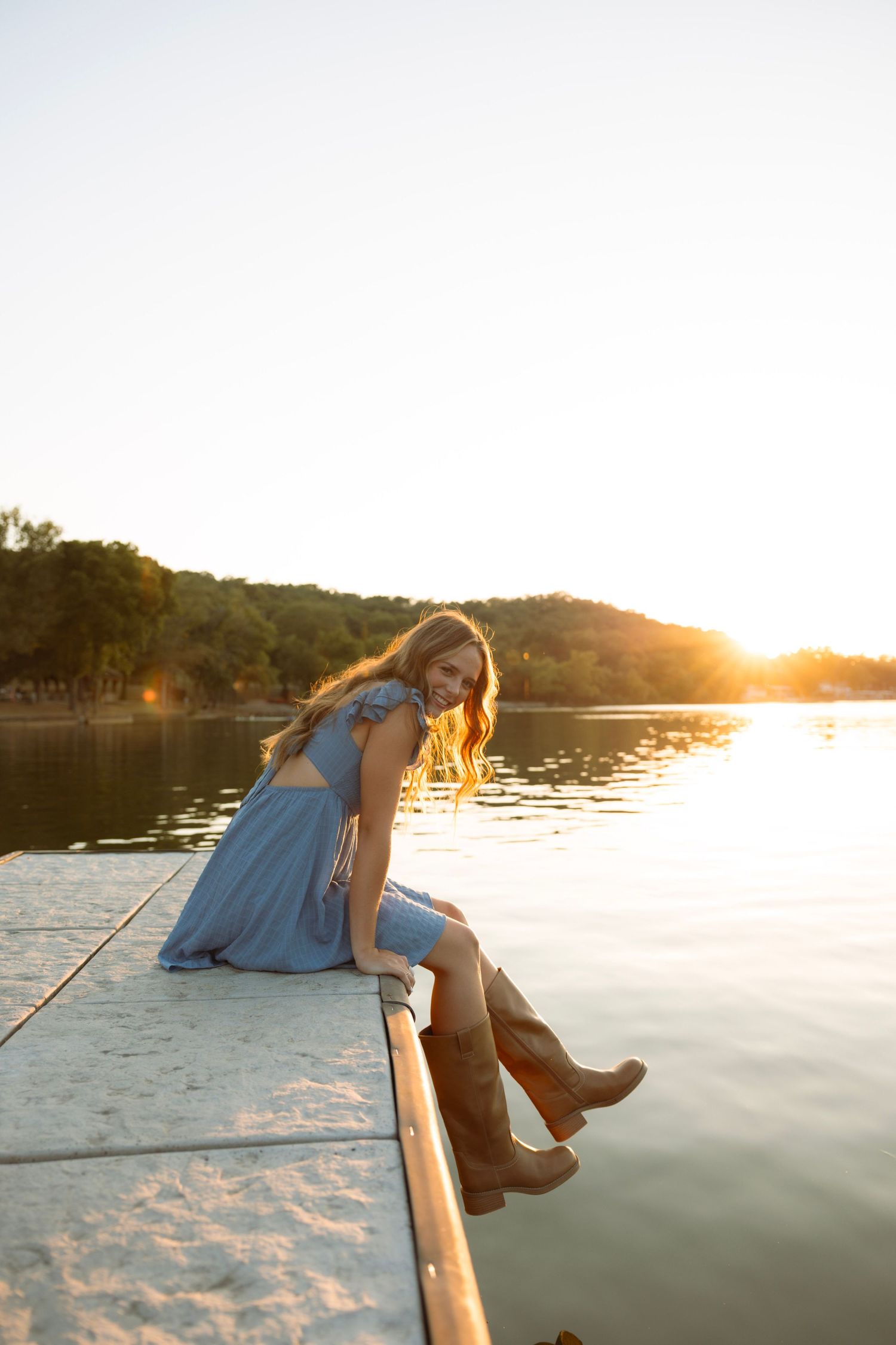 Young woman in blue dress and cowboy boots sits on a dock at golden hour sunset over a calm lake.