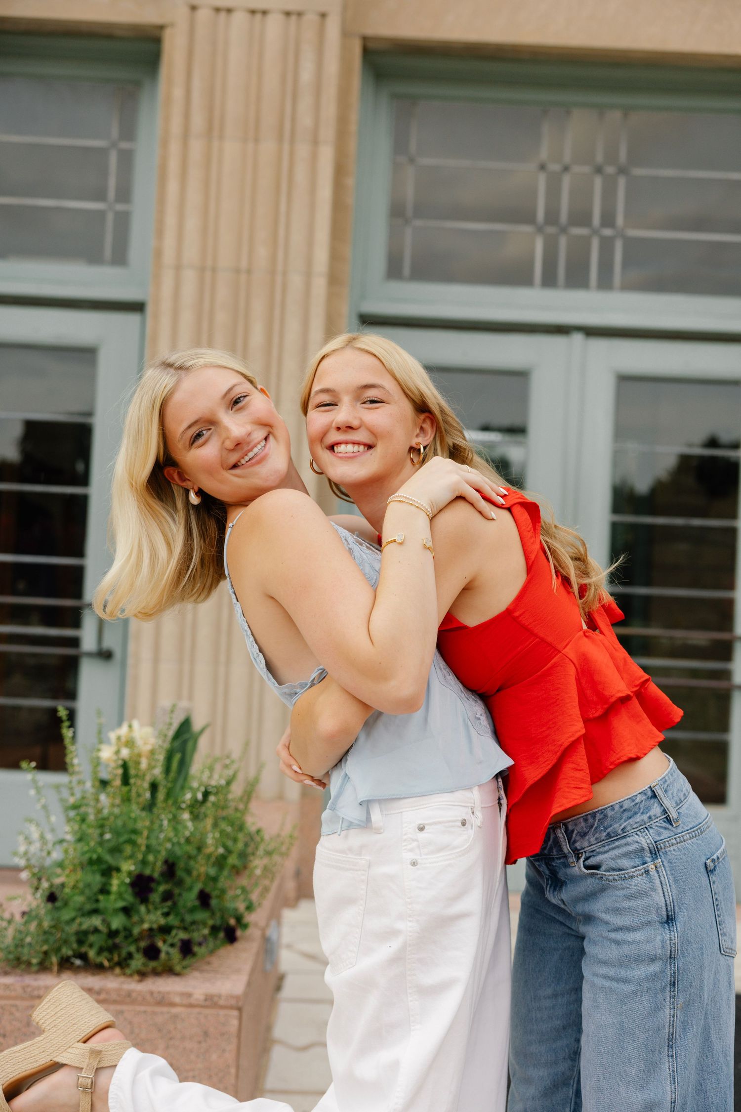 Two smiling blonde women hugging outdoors near a building, one wearing a red top and jeans, the other in a blue top and white skirt.