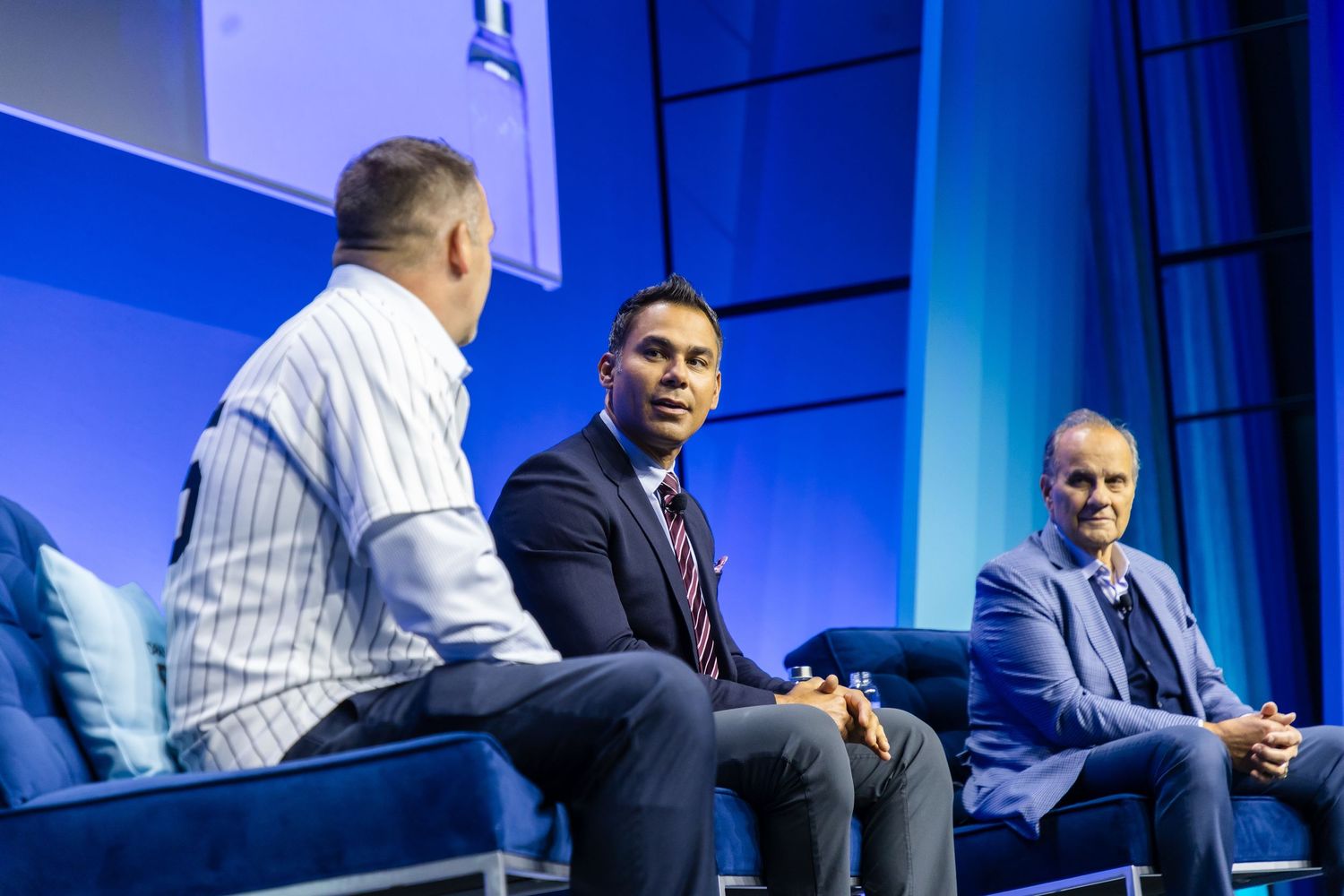 Panel discussion participants engage in conversation on a brightly lit blue stage during a business conference event in New York City.