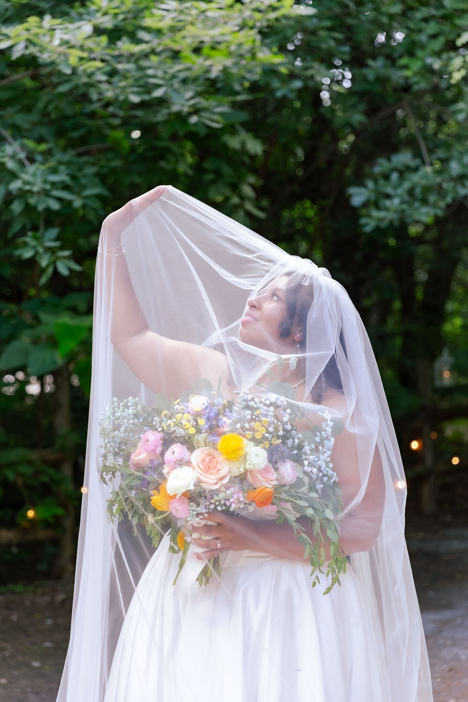 A bride in a white wedding dress and veil holds a vibrant bouquet with orange and pink flowers against a garden backdrop.