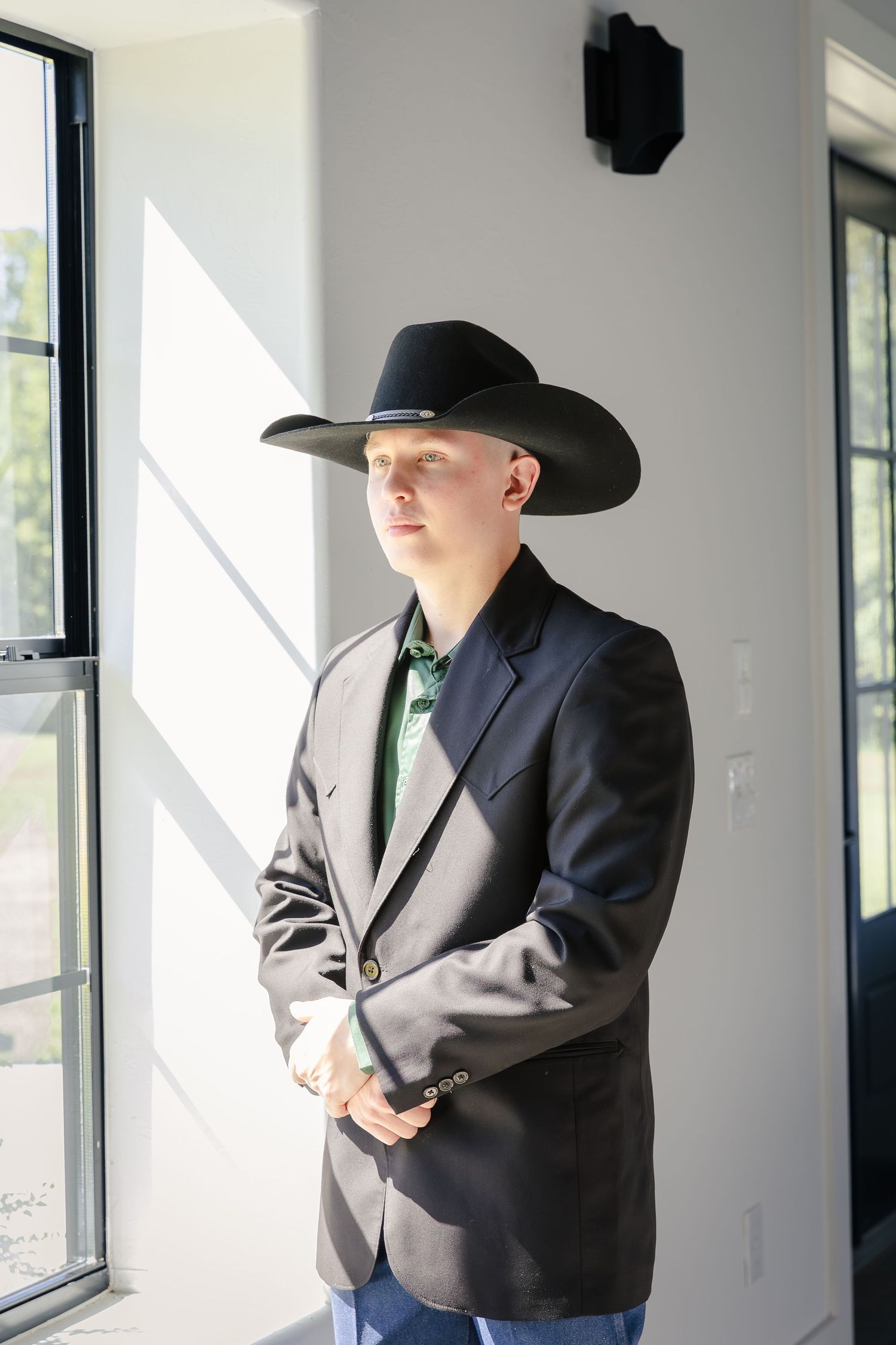 A groom, dressed in a black cowboy hat and dark suit, stands near a window in a minimalist interior space.