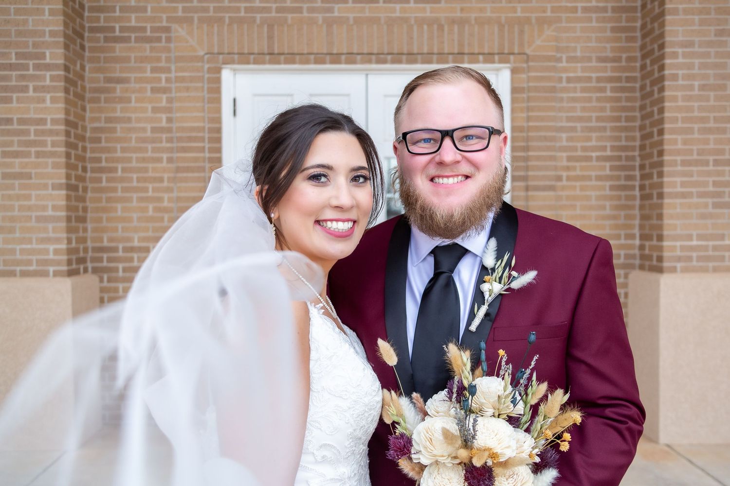 Elegant wedding couple poses in front of a brick wall, the bride in a white dress and the groom in a burgundy suit with a floral bouquet.