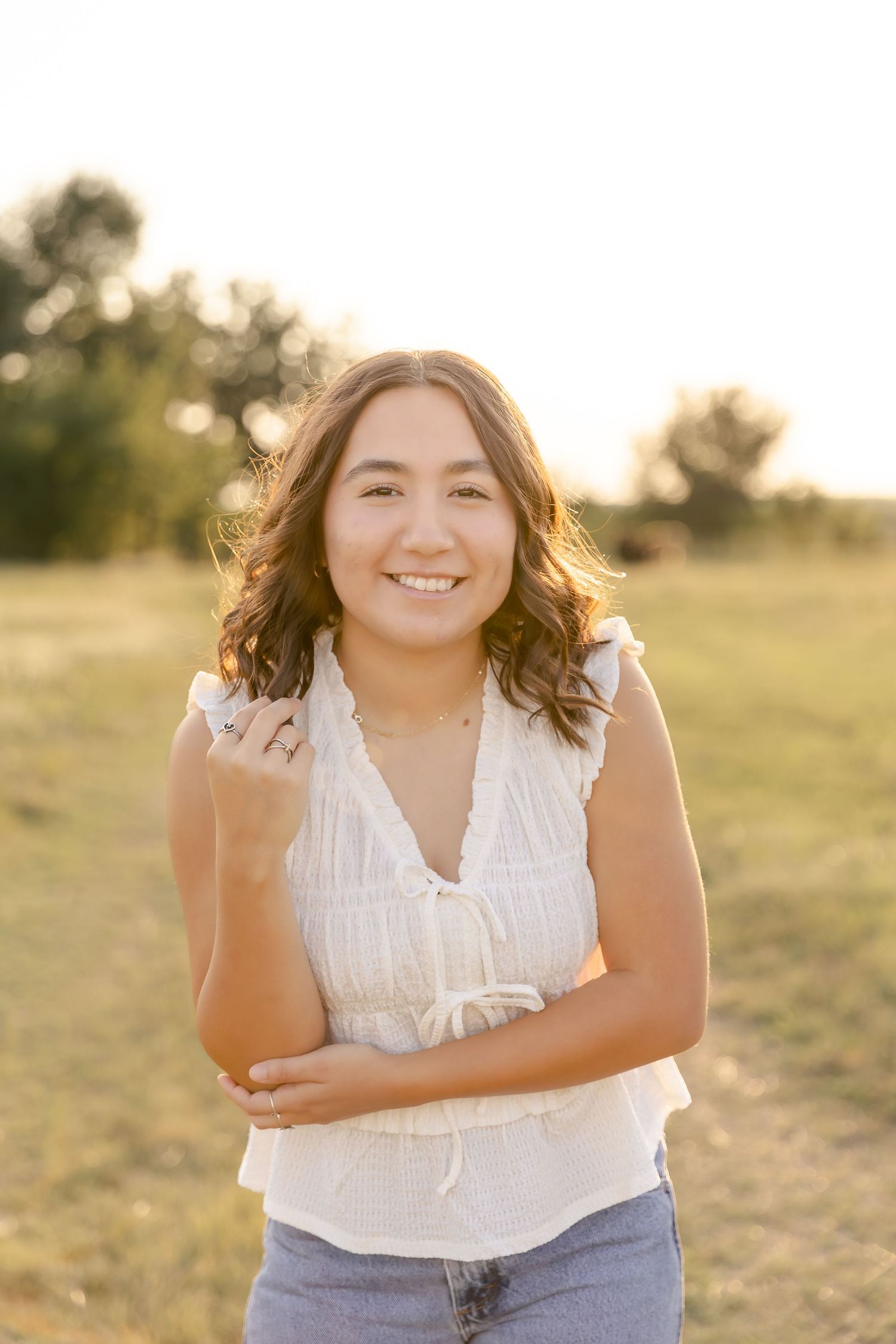 A senior in a white sleeveless top and jeans outfit captured in golden hour lighting on a grassy field.