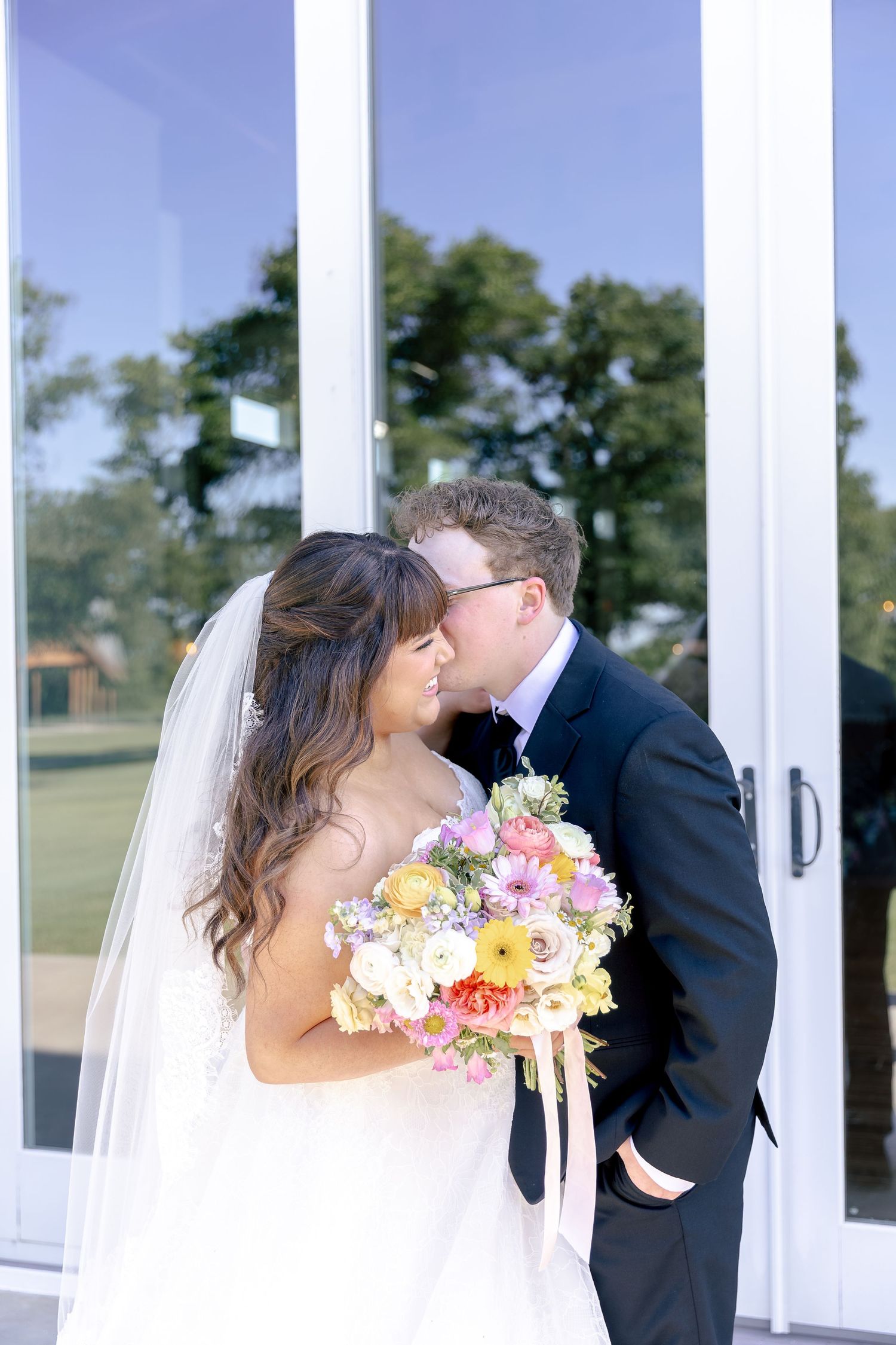 Bride and groom share a tender kiss outdoors, bride holding a colorful bouquet of pink, yellow, and white flowers.
