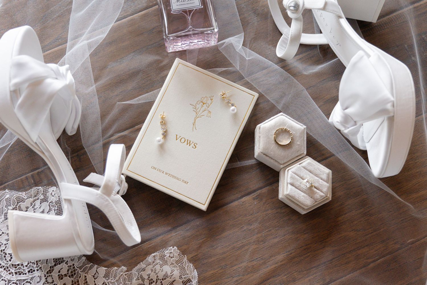 Elegant wedding flat lay with vows book, ring boxes, pearl earrings, bridal heels, and veil on wood surface.
