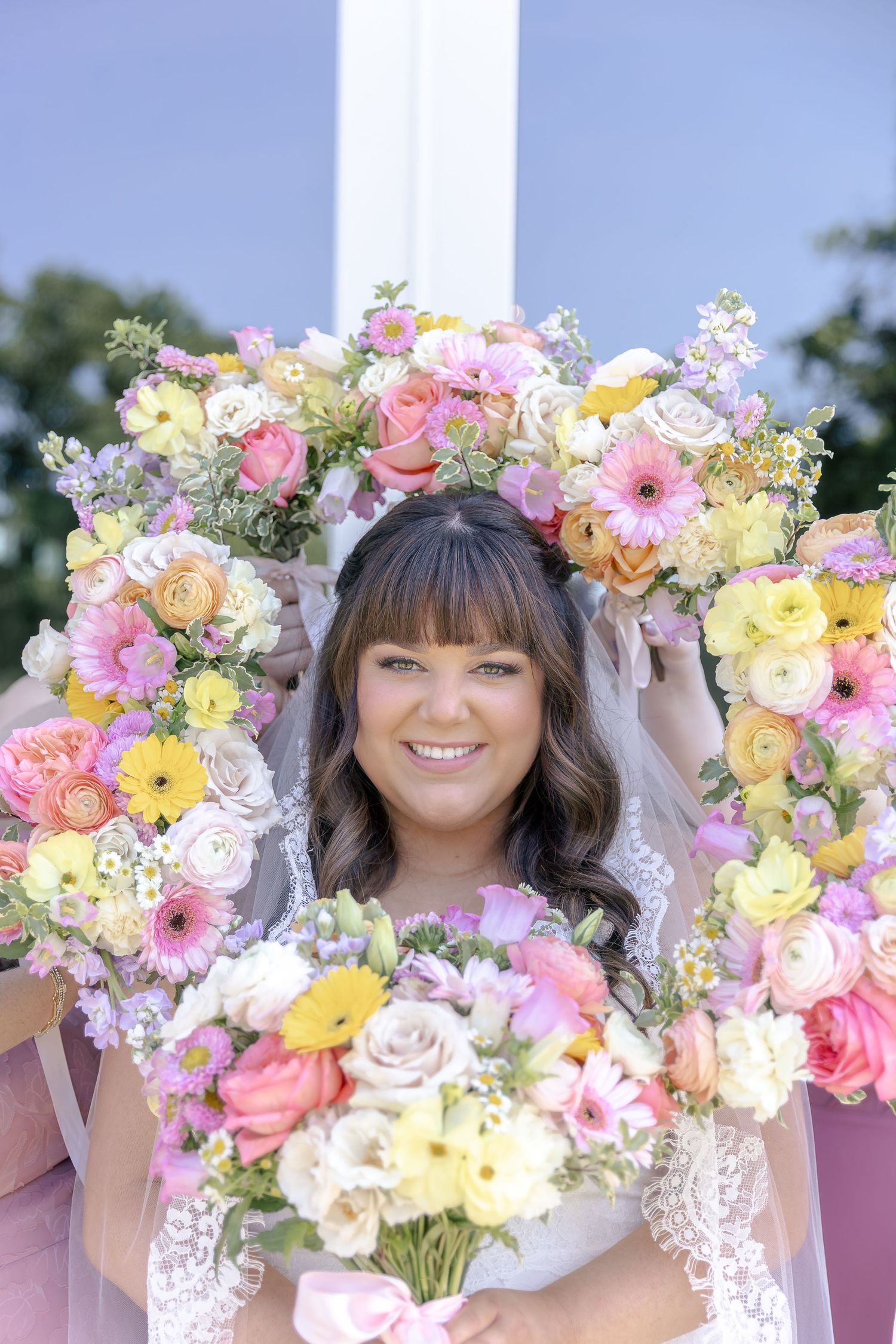 Smiling bride framed by colorful pastel floral arrangements holding a bouquet with pink ribbon on her wedding day.