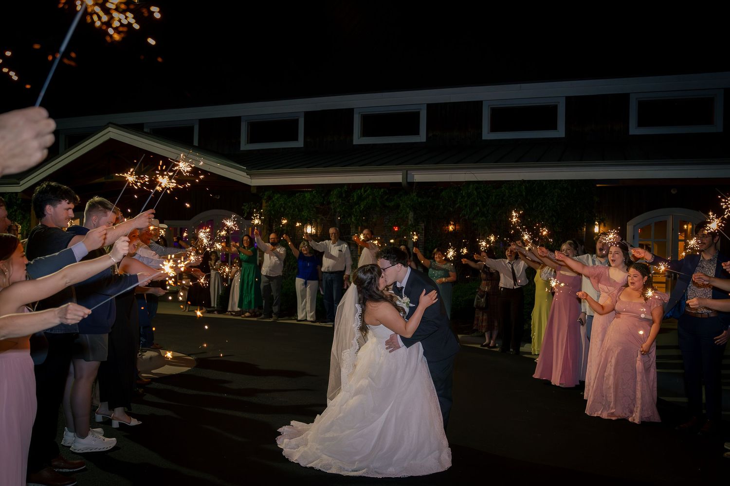 Newlyweds share a kiss during a sparkler send-off at a night wedding reception surrounded by guests.