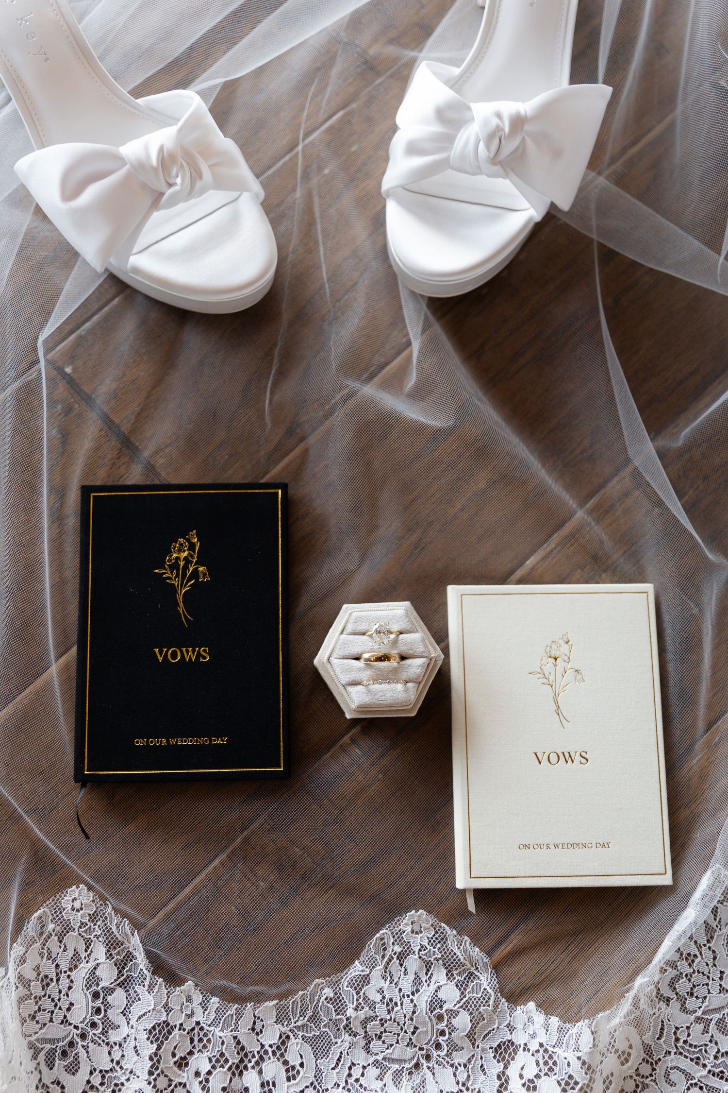 Wedding flat lay with vow books, diamond rings in velvet box, white bow heels, and bridal veil on wood floor.