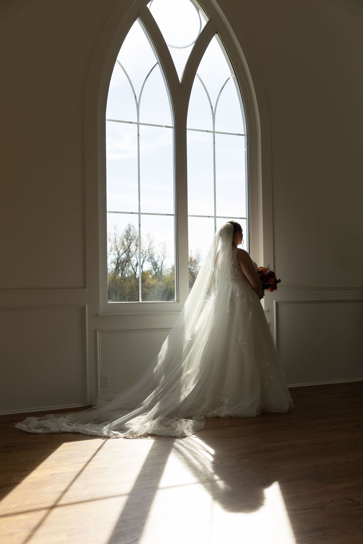 Silhouette of a bride in a flowing white gown against gothic windows in a sunlit chapel.
