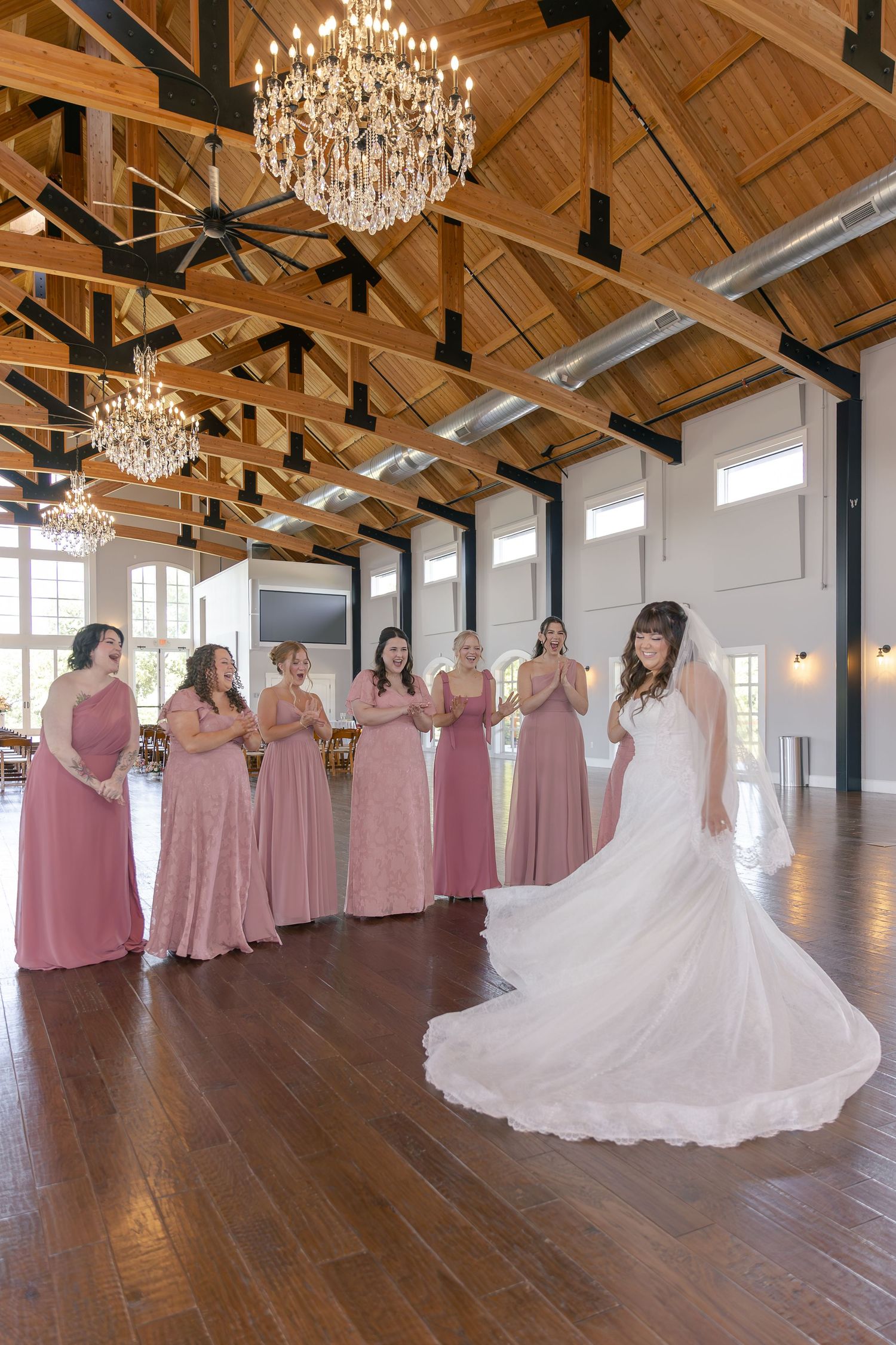 Bride in white gown reveals dress to bridesmaids in pink dresses inside elegant wood-beamed venue with crystal chandeliers.