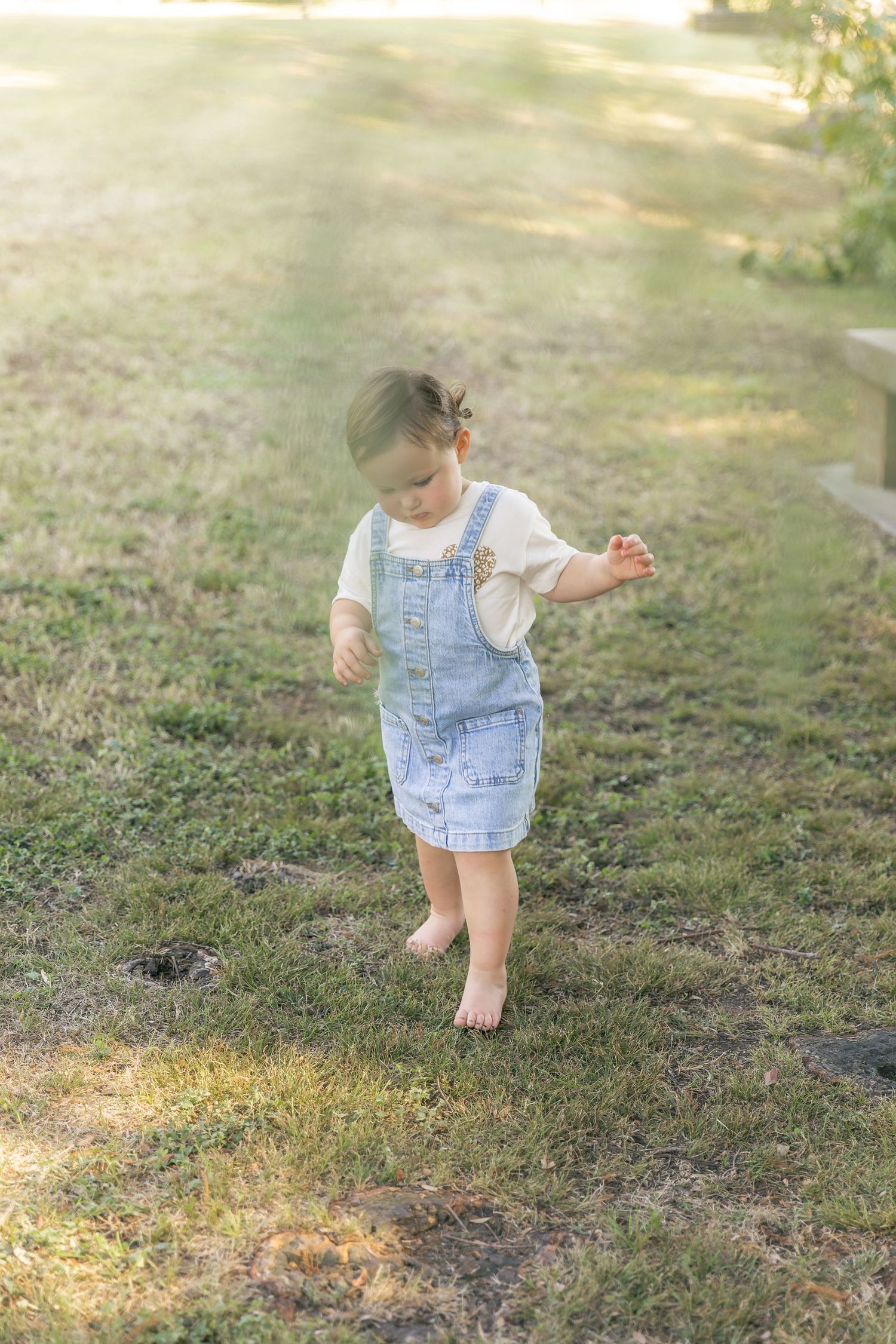 Toddler in denim overalls exploring and walking on grassy ground during photoshoot.