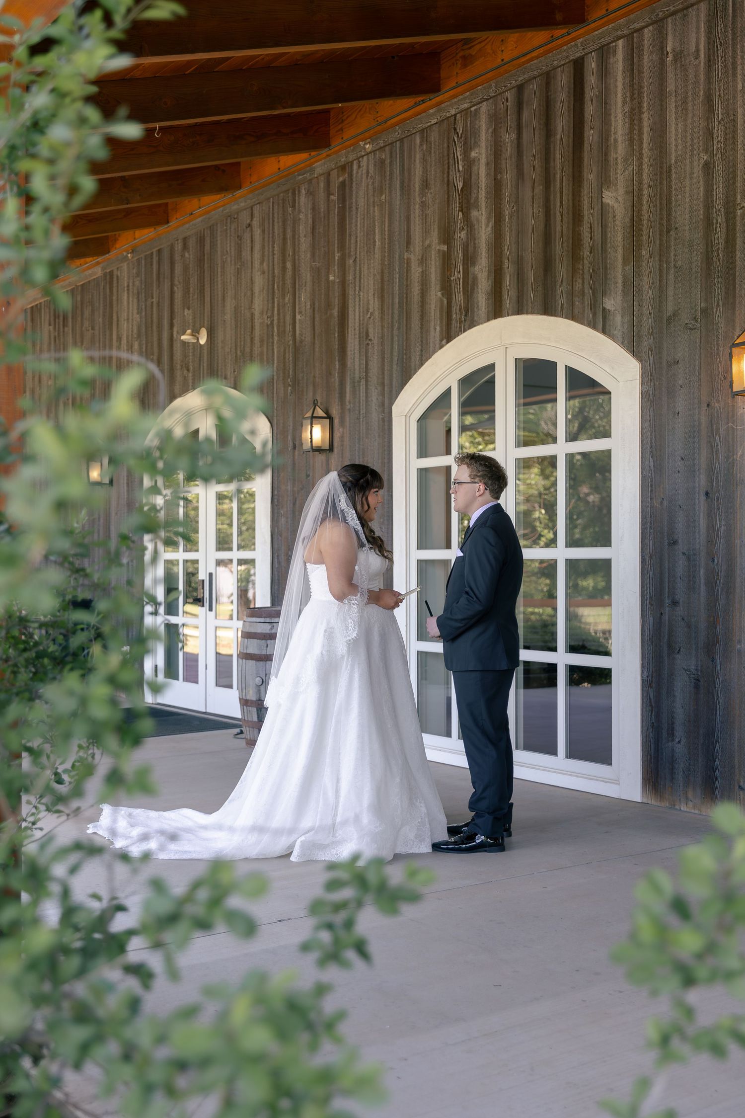 Bride and groom share a tender first look moment on a rustic barn venue porch, surrounded by lush greenery.