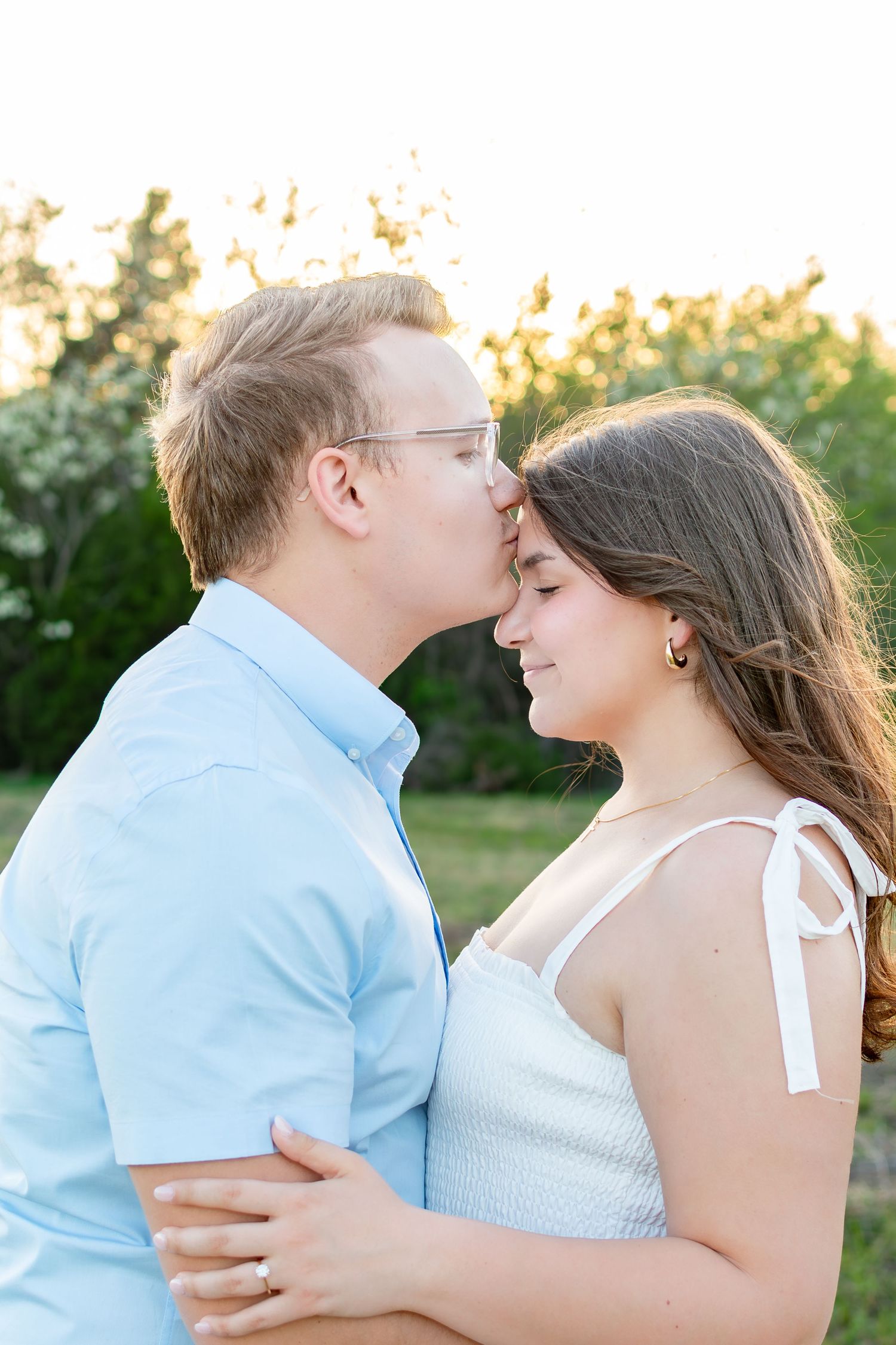 Couple in blue shirt and white dress share intimate moment during sunset engagement shoot.