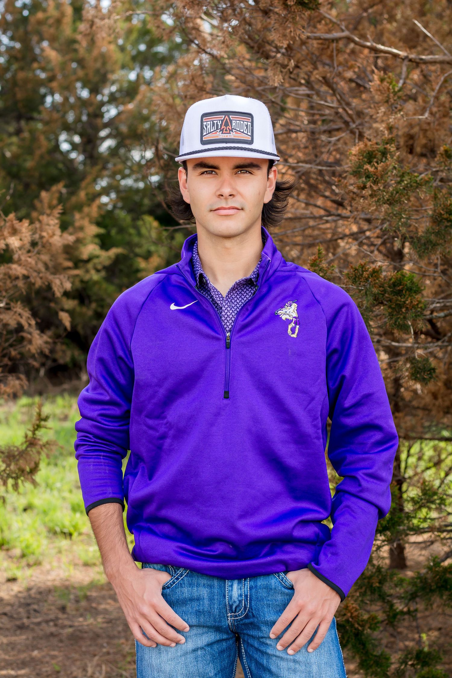 Athletic senior portrait in purple Nike quarter-zip pullover and baseball cap against a natural background.