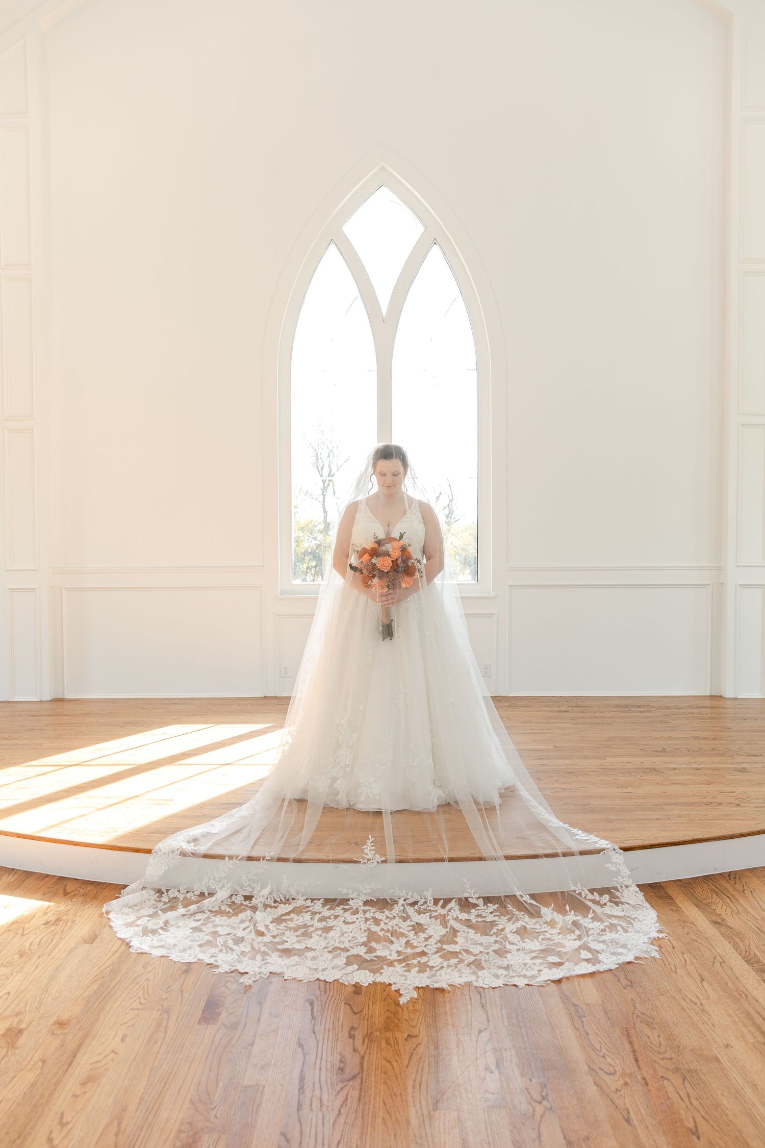 A stunning white wedding dress with a lace train photographed in a bright chapel with arched windows.