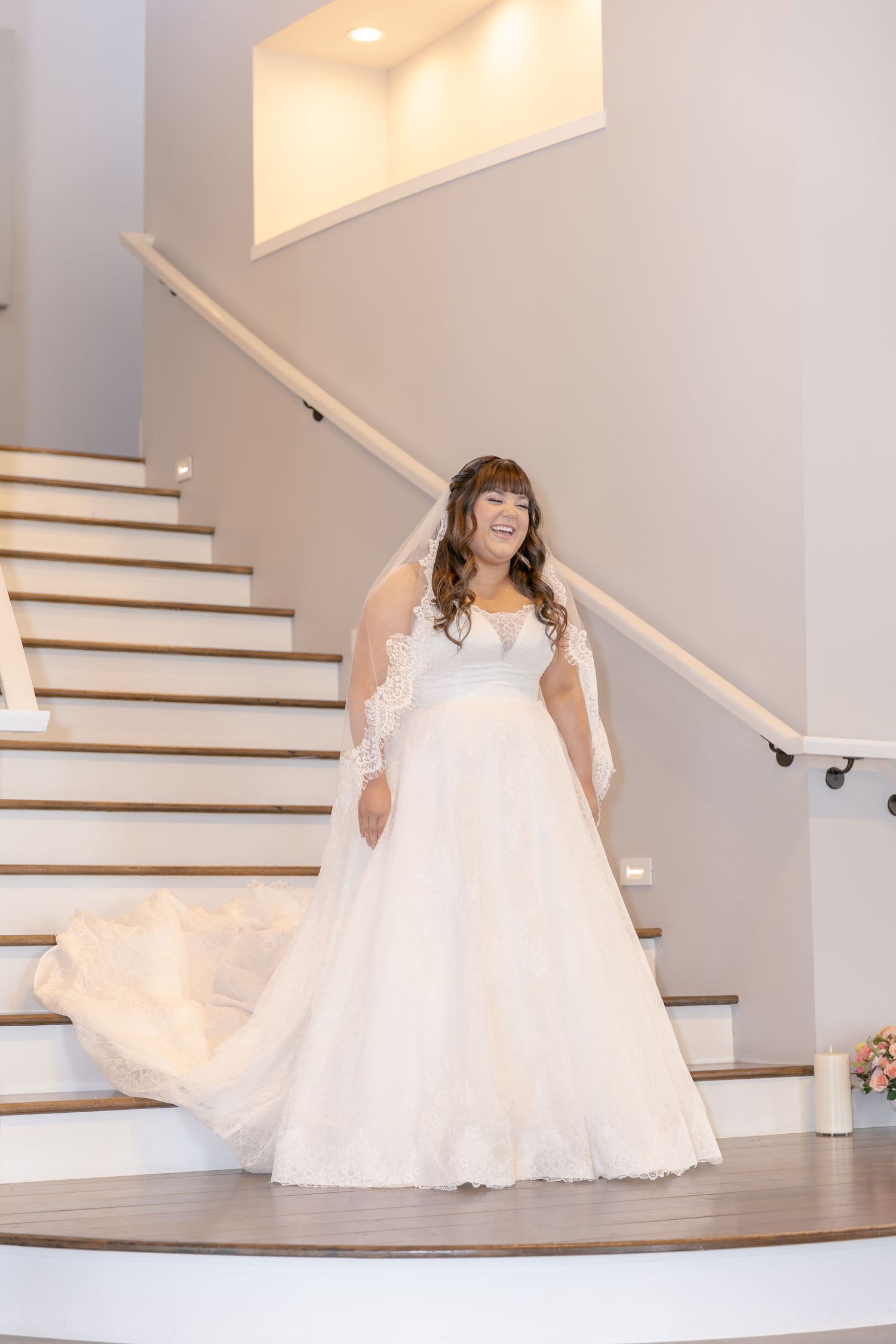 Bride in elegant white lace ball gown and veil poses gracefully on curved staircase indoors.