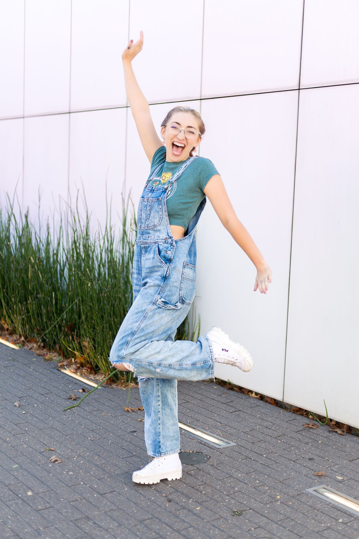 Playful senior poses in denim overalls and green shirt against a white wall with grass.