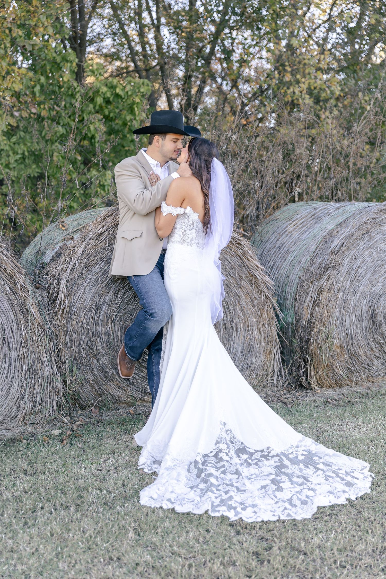 A romantic country wedding photo with a couple embracing by hay bales, featuring a flowing lace gown with a long train.
